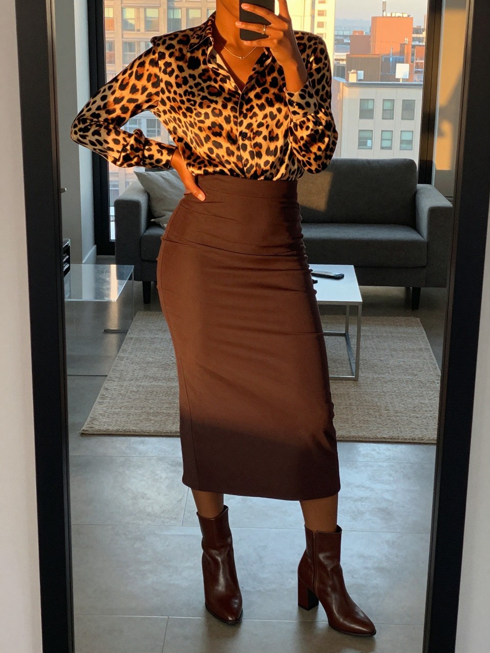 mirror selfie of a black woman wearing a leopard print silk blouse tucked into a chocolate brown midi skirt with brown block-heel ankle boots, one hand on her hip, cropped at NECK so face doesn't show, in a city apartment, evening golden hour, iPhone photo quality.