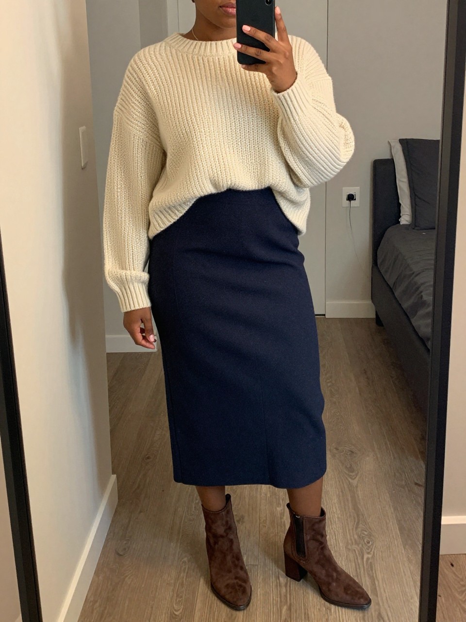mirror selfie of a curvy black woman wearing an oversized cream chunky sweater half-tucked into a navy wool midi skirt with brown suede block-heel ankle boots, one hand holding her phone at her side, cropped at NECK so face doesn't show, in a city apartment, warm moody lighting, iPhone photo quality.