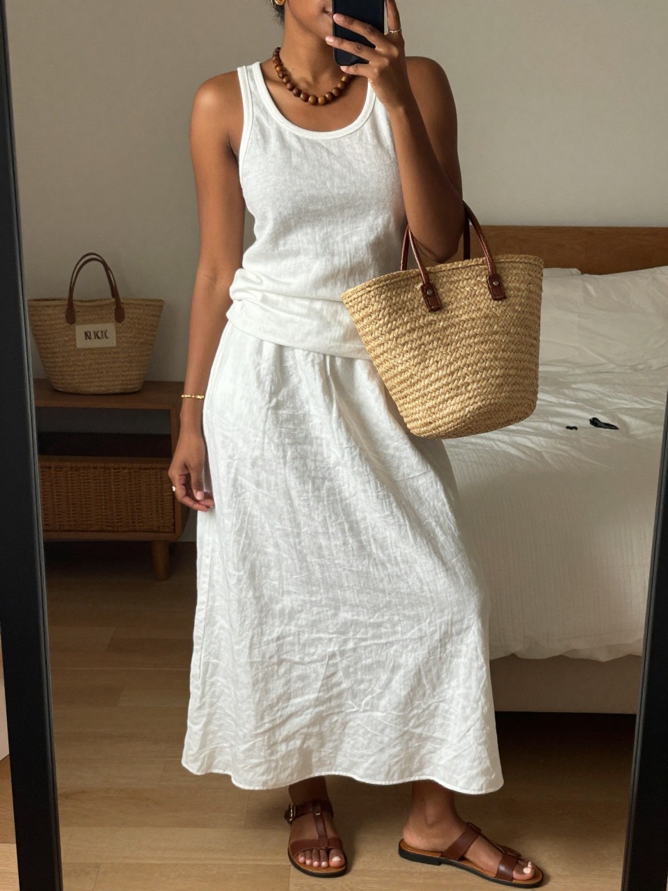 mirror selfie of a black woman wearing a white linen tank top tucked into a white linen maxi skirt with brown leather flat sandals and a large straw basket bag, a wooden bead necklace visible, tortoiseshell sunglasses on, standing in an easy summer pose, cropped at NECK so face doesn't show, in a feminine-themed bedroom, warm moody lighting, iPhone photo quality.