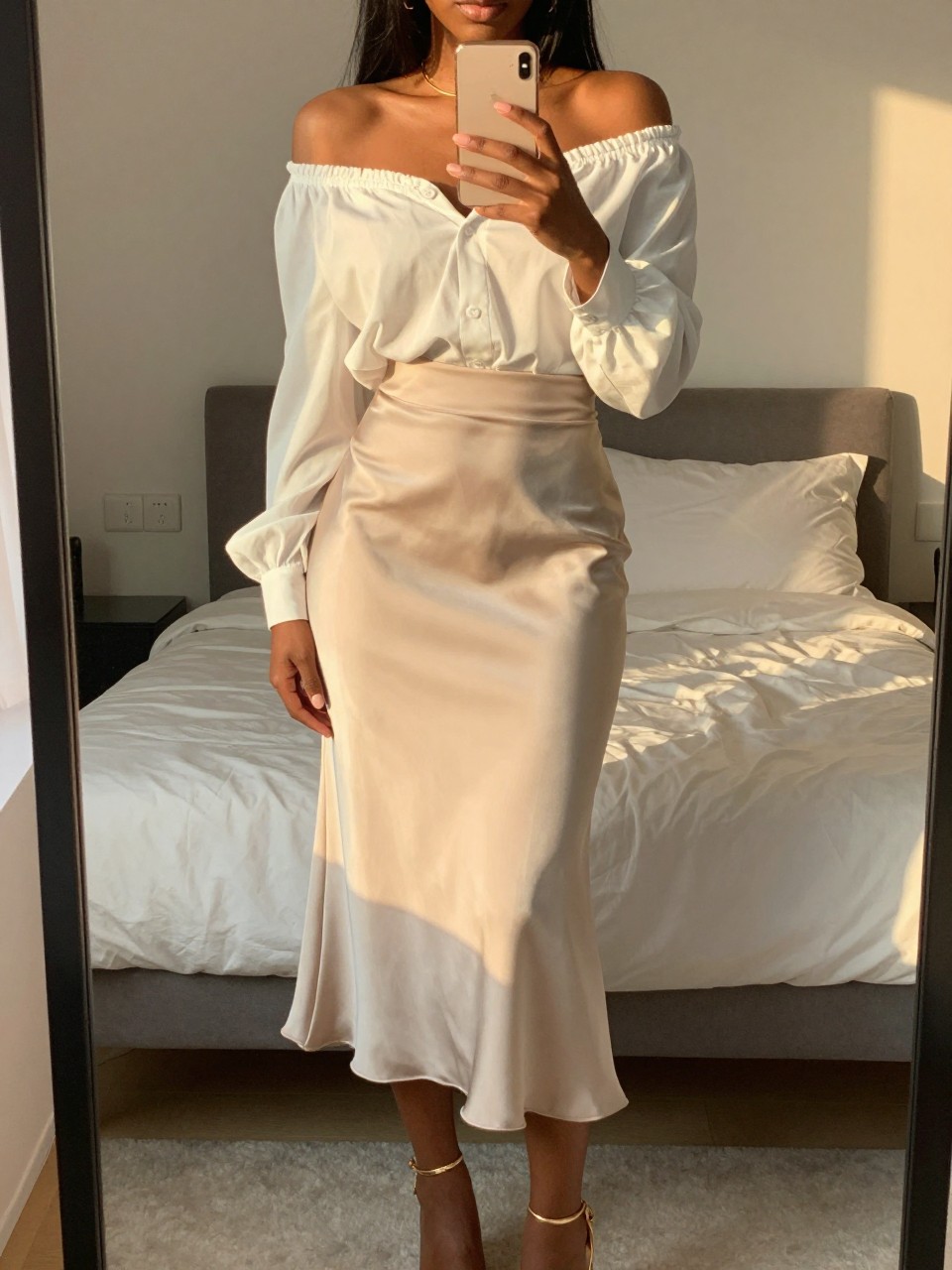 iPhone mirror selfie of a black woman wearing a white off-the-shoulder blouse tucked into a soft cream satin midi skirt with gold strappy heels, one hand holding her phone at chest level, cropped at NECK so face doesn't show, in a feminine-themed bedroom, evening golden hour, iPhone photo quality.