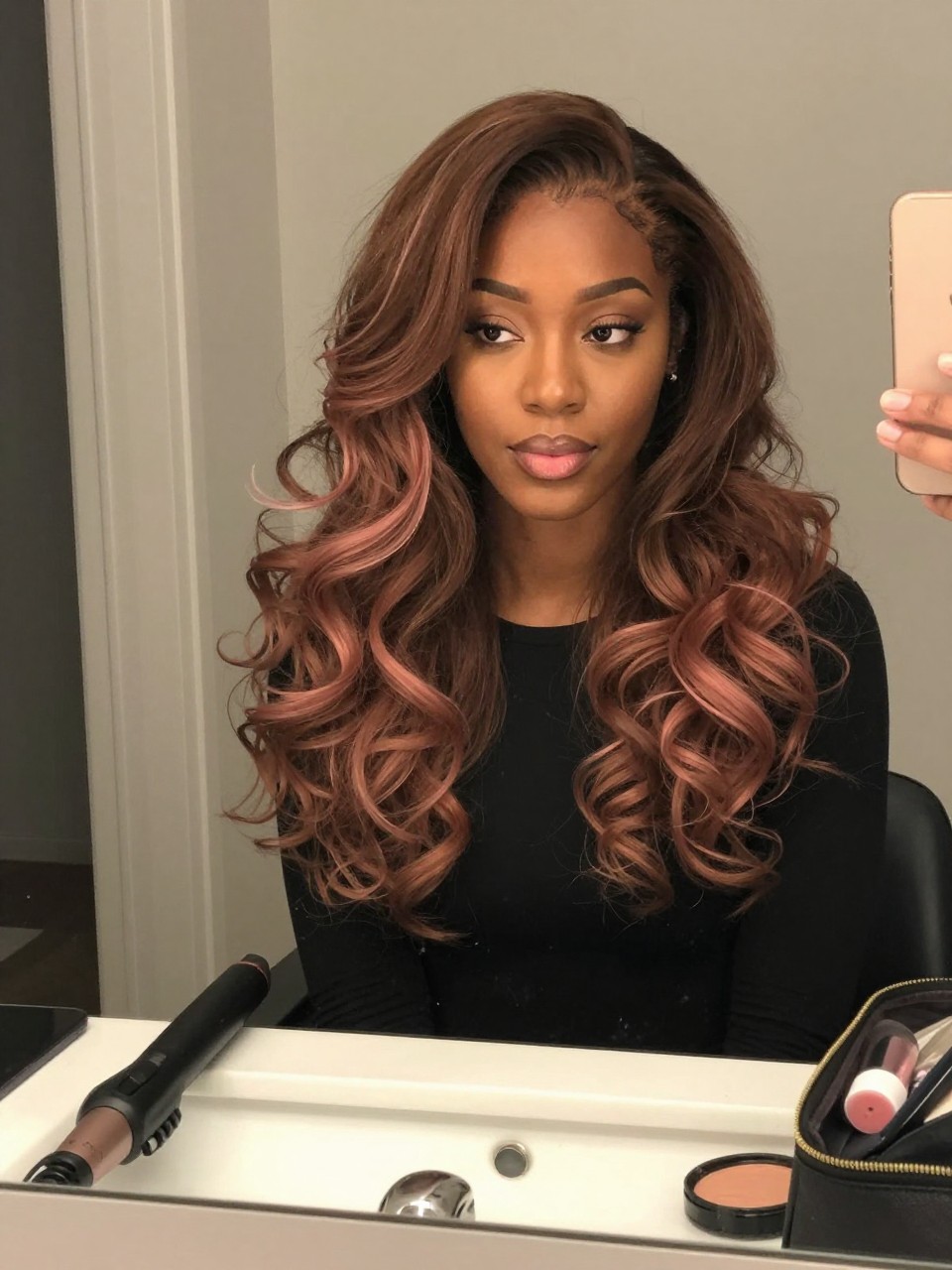 Photo of a black woman wearing soft brown hair with pastel rose brown tips in loose curls, candid mirror selfie, bathroom vanity mirror setting, soft indoor lighting, with a curling wand on the counter and a makeup bag nearby, latest iPhone photo quality.