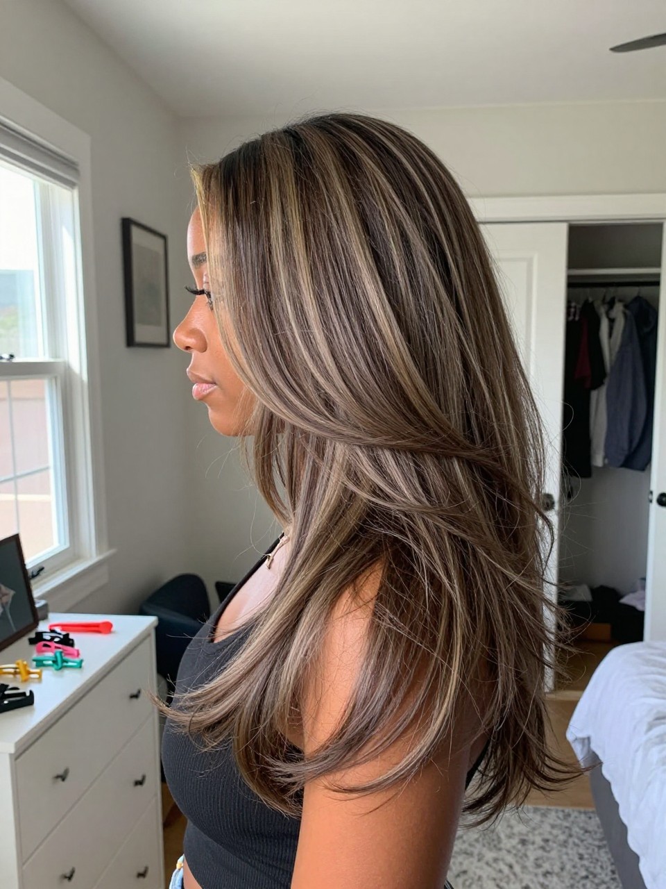 Photo of a black woman wearing long layered ash brown hair with mushroom brown highlights and soft movement, side profile view, in a sun-drenched bedroom, natural window light, containing a small lived-in detail such as sectioning clips on the dresser and a slightly open closet behind, latest iPhone photo quality.