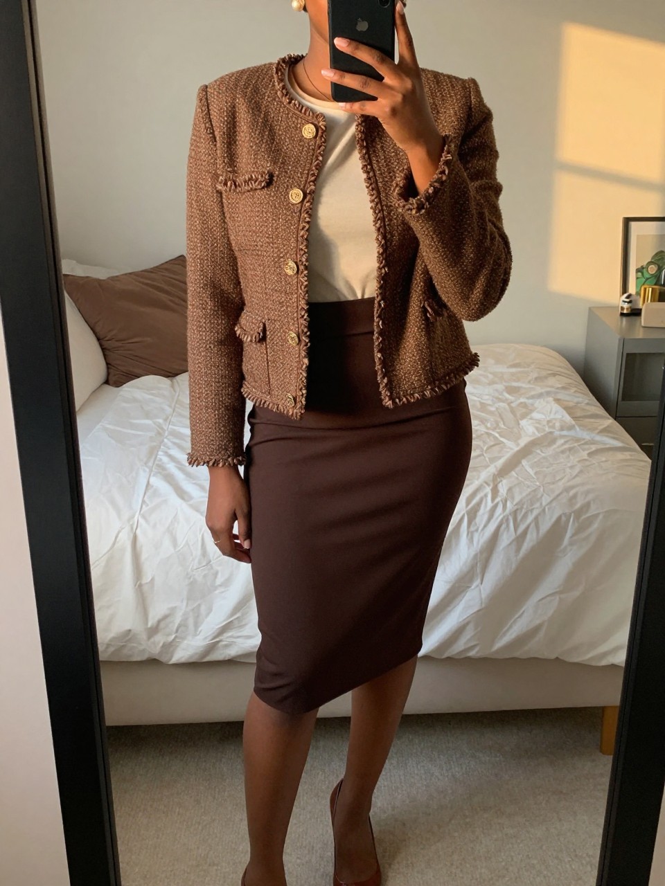 mirror selfie of a black woman wearing a brown tweed jacket over a cream shell tucked into a chocolate brown pencil skirt with pearl earrings and brown pumps, one hand resting on her jacket, cropped at NECK so face doesn't show, in a feminine-themed bedroom, evening golden hour, iPhone photo quality.