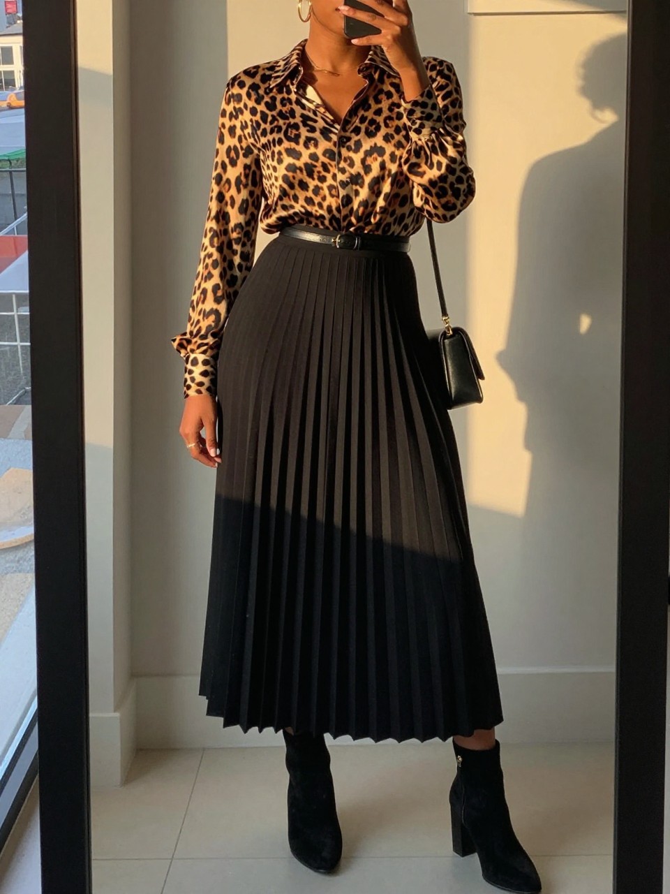 mirror selfie of a black woman wearing a leopard print silk blouse tucked into a black pleated maxi skirt with black suede block-heel ankle boots and a thin black belt, gold hoop earrings visible, holding a black crossbody bag, standing in a confident pose, cropped at NECK so face doesn't show, in a city apartment, evening golden hour, iPhone photo quality.