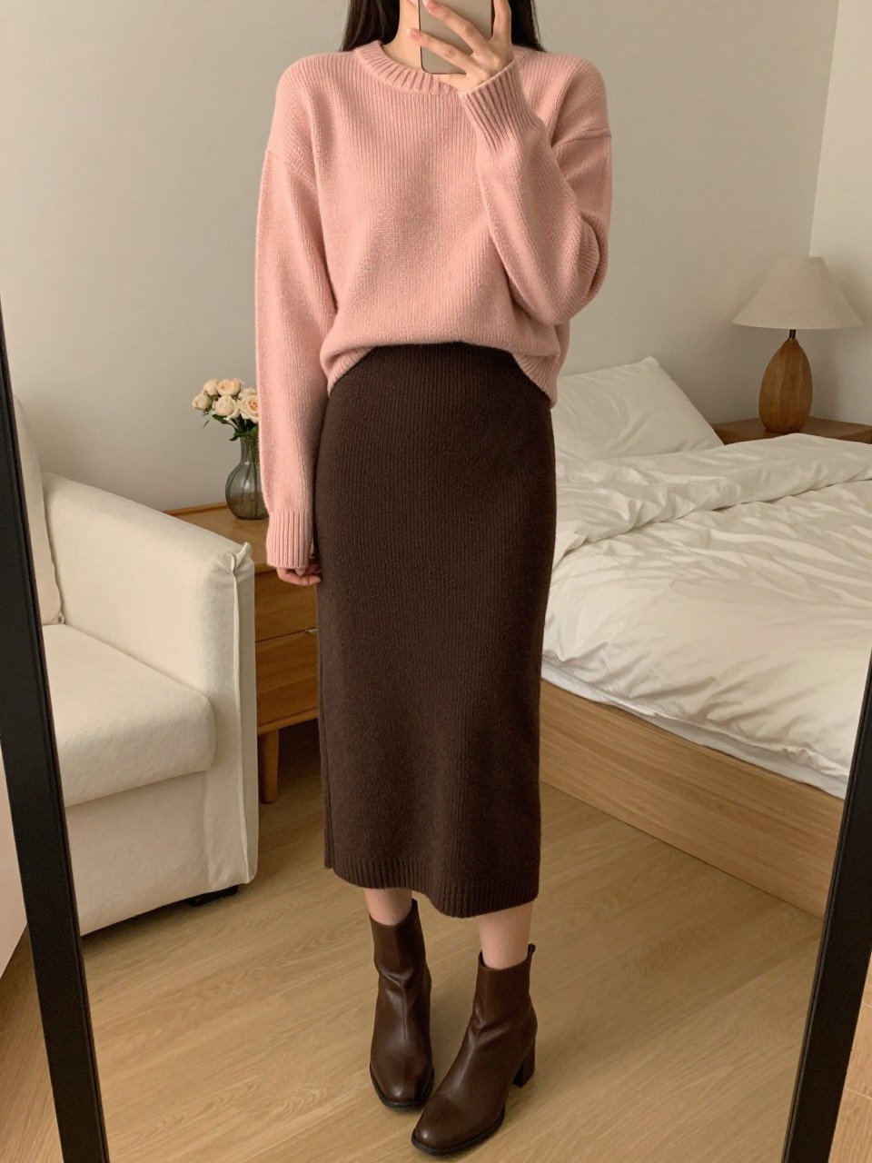 iPhone mirror selfie of a woman wearing a blush pink crewneck sweater tucked into a chocolate brown knit midi skirt with brown block-heel ankle boots, one hand adjusting her sleeve, cropped at NECK so face doesn't show, in a feminine-themed bedroom, warm moody lighting, iPhone photo quality.