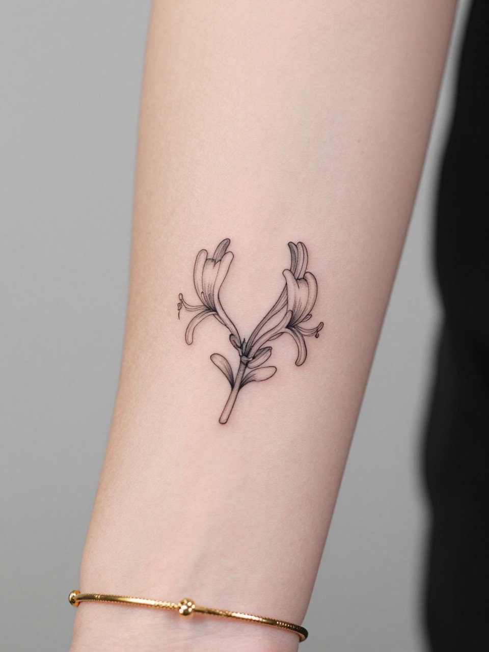 Image Prompt: "Macro shot of a tiny honeysuckle flower tattoo on the inner wrist, black ink fine line floral design with small trumpet-shaped blooms and light shading, minimalist style, realistic skin texture, delicate gold bracelet, tight crop, feminine tattoo inspiration image, clean minimal background, tattoo-centered composition (no watermarks on images)"