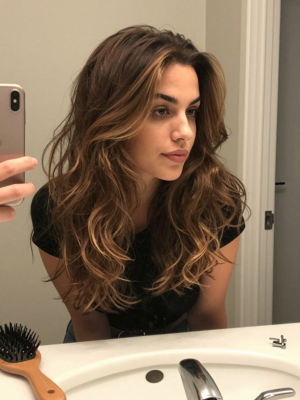 Photo of a woman wearing a wavy brown lob with mushroom highlights and soft lowlights, candid mirror selfie, at a bathroom vanity mirror, soft indoor lighting, containing a small lived-in detail such as a hairbrush on the sink and a silver clip nearby, latest iPhone photo quality.