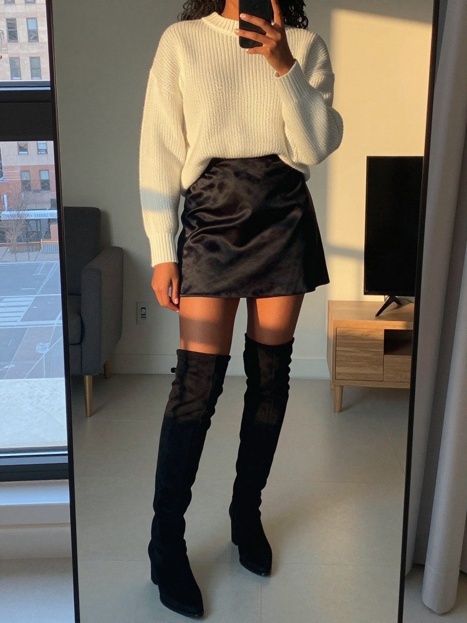 iPhone mirror selfie of a black woman in a white knit sweater and black satin skirt, pulling on over-the-knee boots, cropped at NECK so face doesn't show, in a city apartment, evening golden hour, iPhone photo quality.