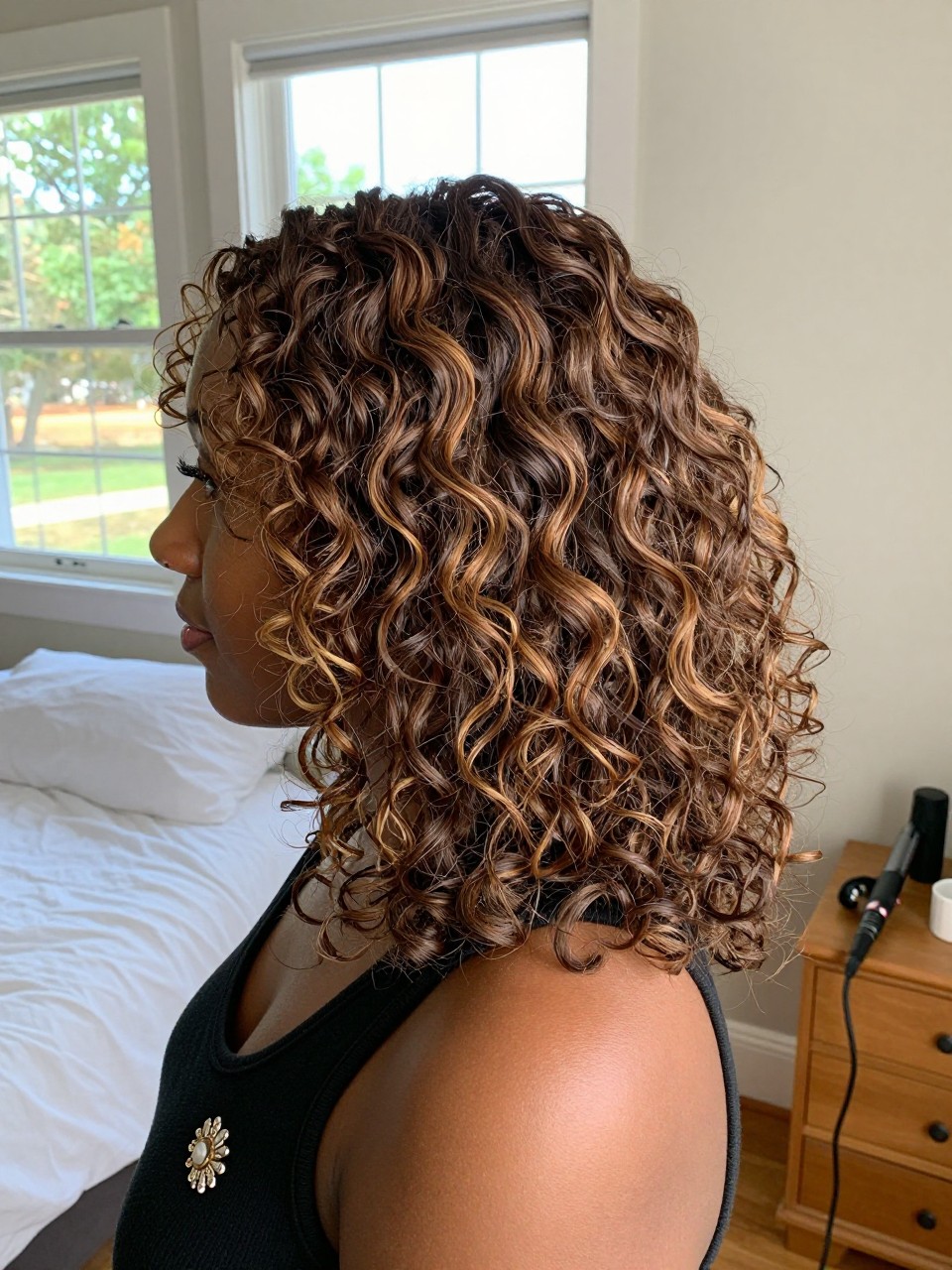 Photo of a brown woman wearing soft brunette curls with caramel blonde highlights, side profile view, in a sun-drenched bedroom, natural window light, containing a small lived-in detail such as a decorative pin on one side and a curling iron resting on the nightstand, latest iPhone photo quality.