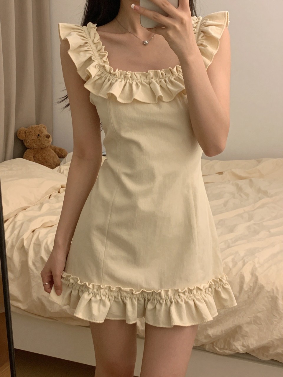 mirror selfie of a woman in a ruffle-trim dress, showing off the detailed hem, cropped at NECK so face doesn't show, in a feminine-themed bedroom, warm moody lighting, iPhone photo quality.