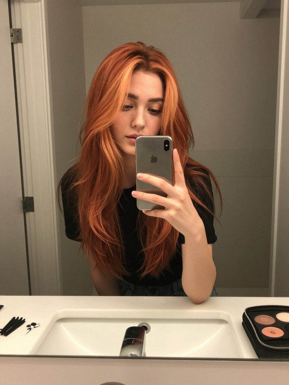 Photo of a woman wearing ginger curtain layers with golden peach highlights around the face, candid mirror selfie, at a bathroom vanity mirror, soft indoor lighting, containing a small lived-in detail such as mini sectioning clips on the counter and a makeup pouch nearby, latest iPhone photo quality.