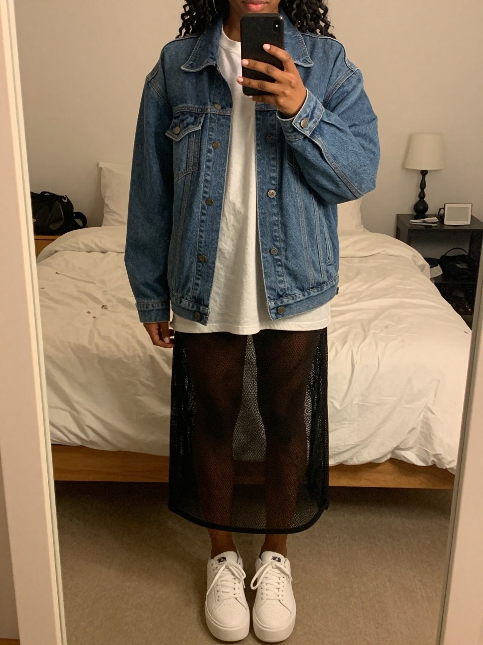 mirror selfie of a black woman wearing an oversized band tee with a blue denim jacket and a black sheer mesh skirt, white platform sneakers, one hand holding her phone low, cropped at NECK so face doesn't show, in a feminine-themed bedroom, warm moody lighting, iPhone photo quality.