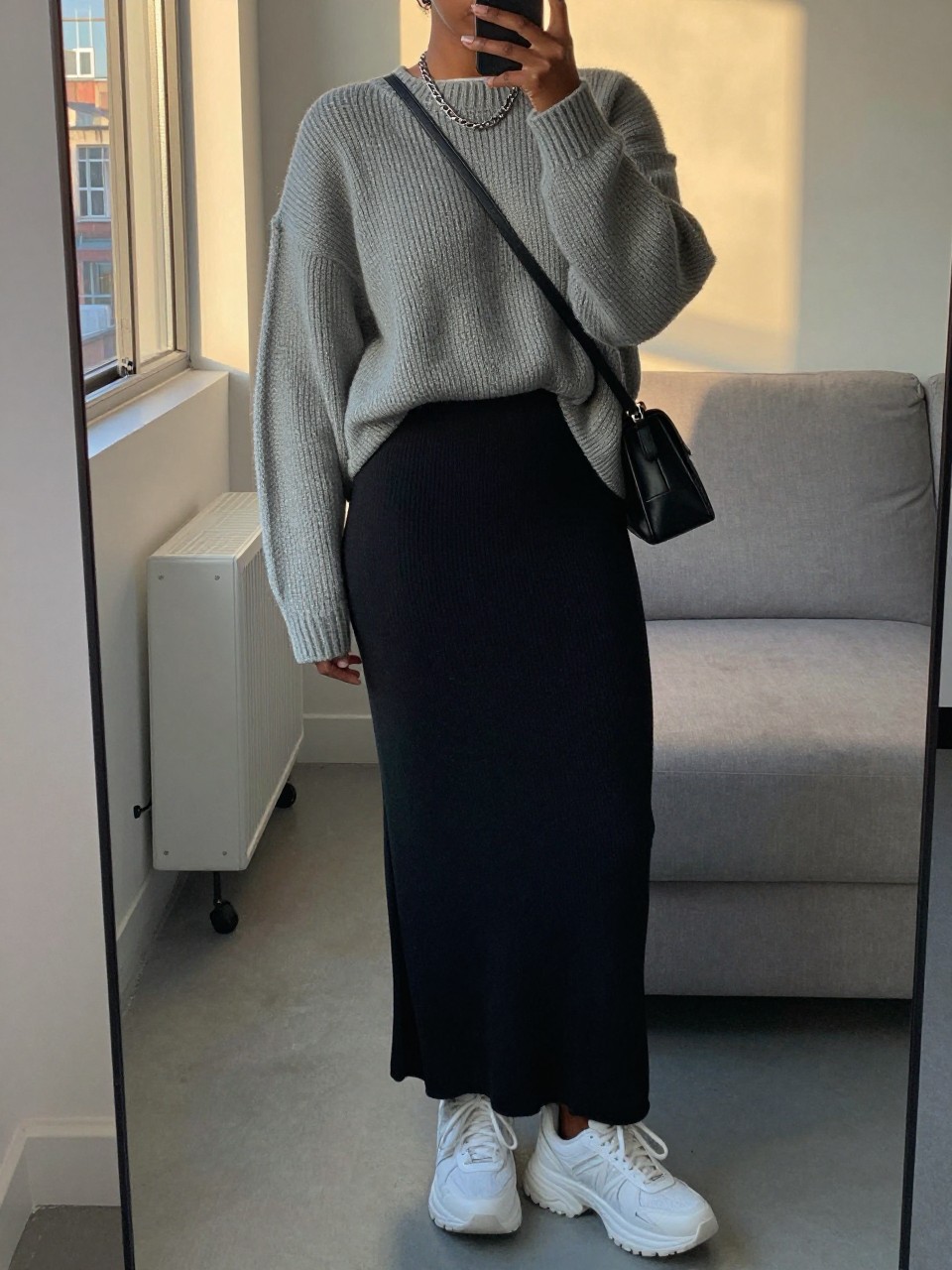 mirror selfie of a black woman wearing an oversized heather grey chunky sweater loosely tucked into a black jersey knit maxi skirt with chunky white sneakers and a silver chain necklace, holding a black crossbody bag, standing in a relaxed cool-girl pose, cropped at NECK so face doesn't show, in a city apartment, evening golden hour, iPhone photo quality.
