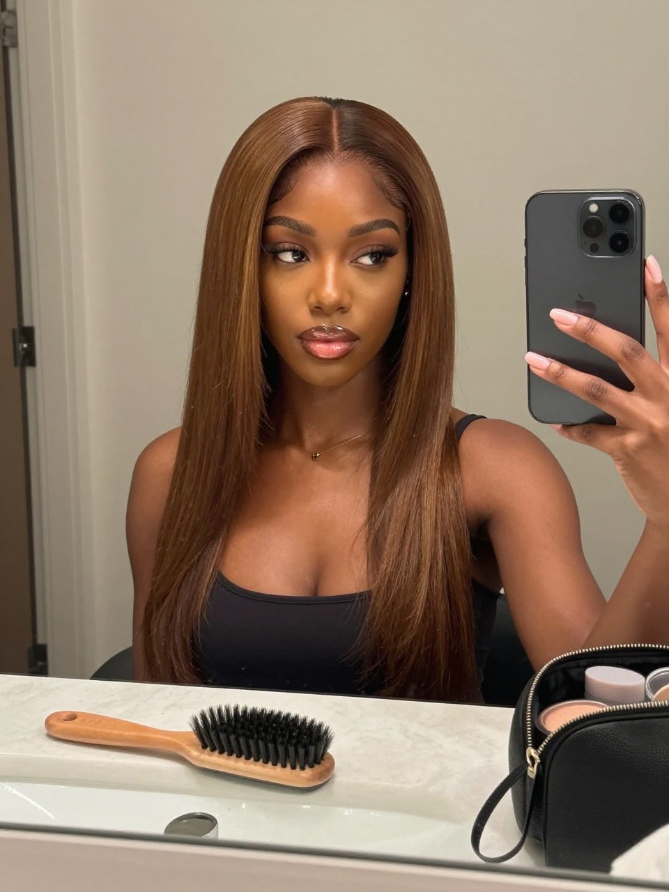 Photo of a black woman wearing light mocha brown glossy hair styled smooth and soft, candid mirror selfie, bathroom vanity mirror setting, soft indoor lighting, with a paddle brush on the counter and a makeup bag nearby, latest iPhone photo quality.