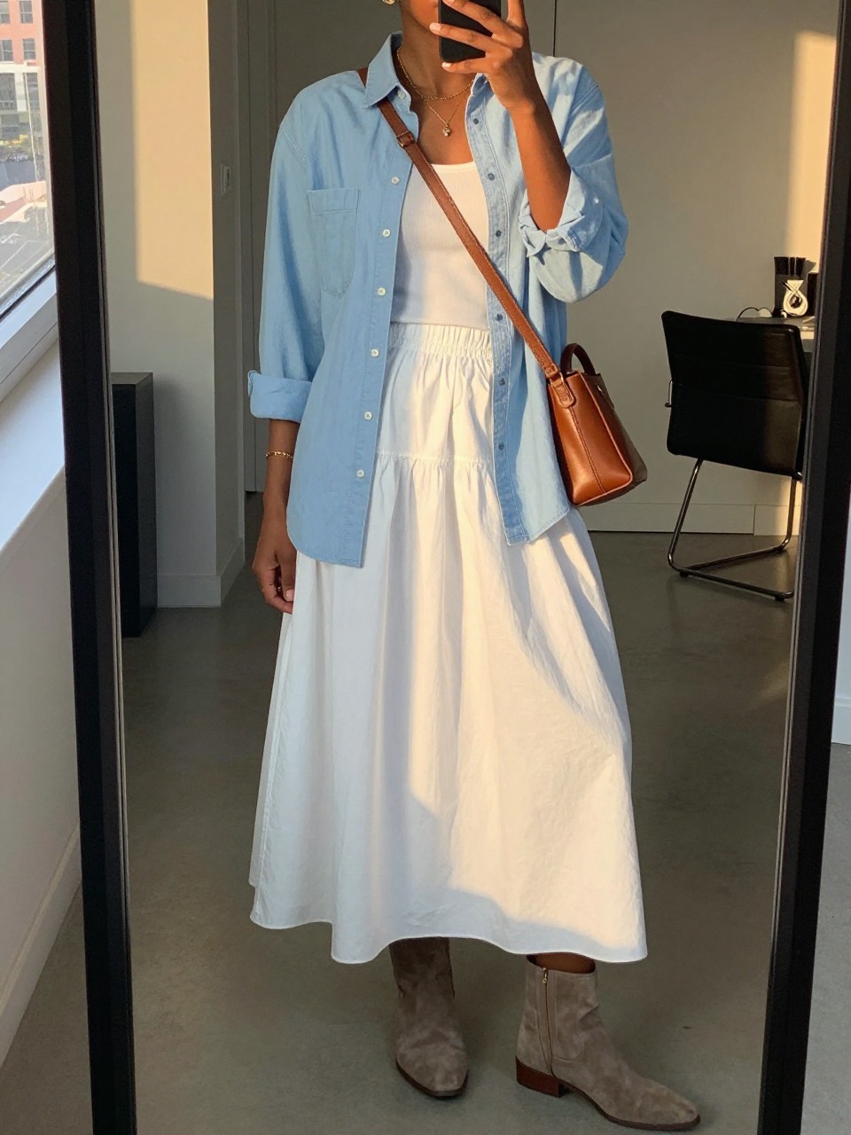 mirror selfie of a black woman wearing a light blue chambray shirt open over a white tank top tucked into a white cotton maxi skirt with flat taupe suede ankle boots and a brown leather crossbody bag, standing in a relaxed layered pose, cropped at NECK so face doesn't show, in a city apartment, evening golden hour, iPhone photo quality.