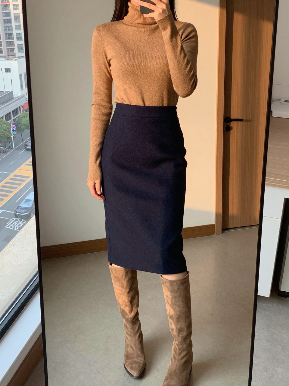 iPhone mirror selfie of a woman wearing a camel turtleneck tucked into a navy wool pencil skirt with camel suede knee-high boots, one hand adjusting her collar, cropped at NECK so face doesn't show, in a city apartment, evening golden hour, iPhone photo quality.