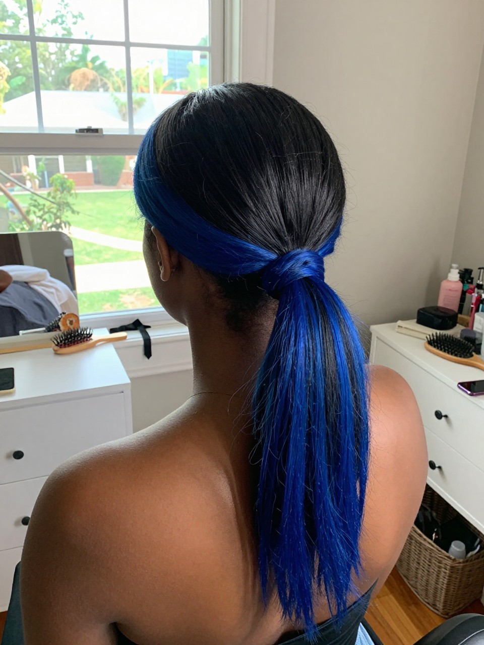 Photo of a black woman wearing a sleek low ponytail with black hair and royal blue highlights through the tail and ends, back-of-head view to highlight details, in a sun-drenched bedroom, natural window light, containing a small lived-in detail such as a black ribbon tied at the base and a hairbrush on the vanity, latest iPhone photo quality.