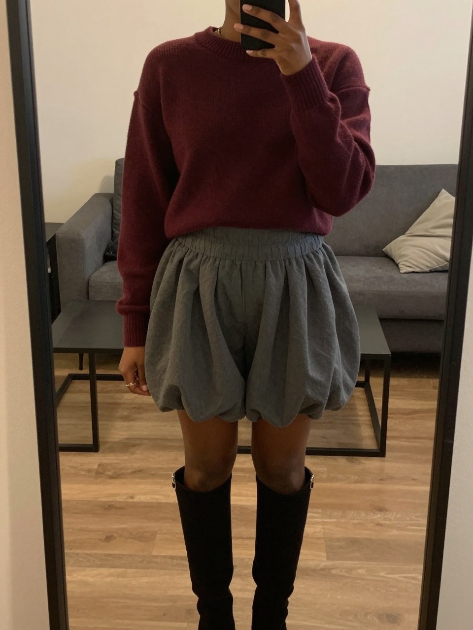 mirror selfie of a black woman in a burgundy cropped sweater and a grey balloon skirt with knee-high boots, standing with phone in hand, cropped at NECK so face doesn't show, in a city apartment, warm moody lighting, iPhone photo quality.