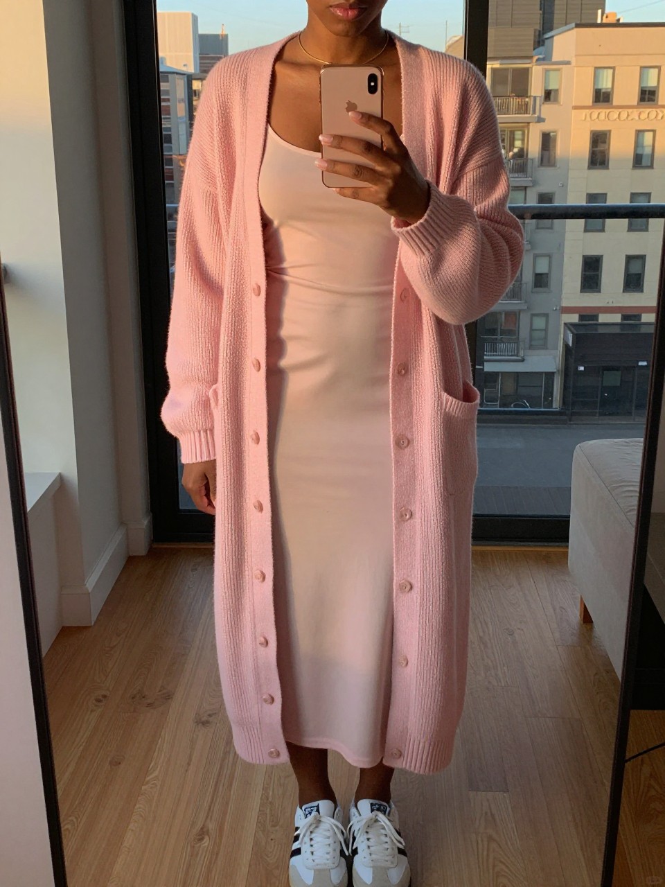 iPhone mirror selfie of a black woman in a long pink cardigan over a tank and slip skirt with sneakers, phone held at mid-chest, cropped at NECK so face doesn't show, in a city apartment, evening golden hour, iPhone photo quality.
