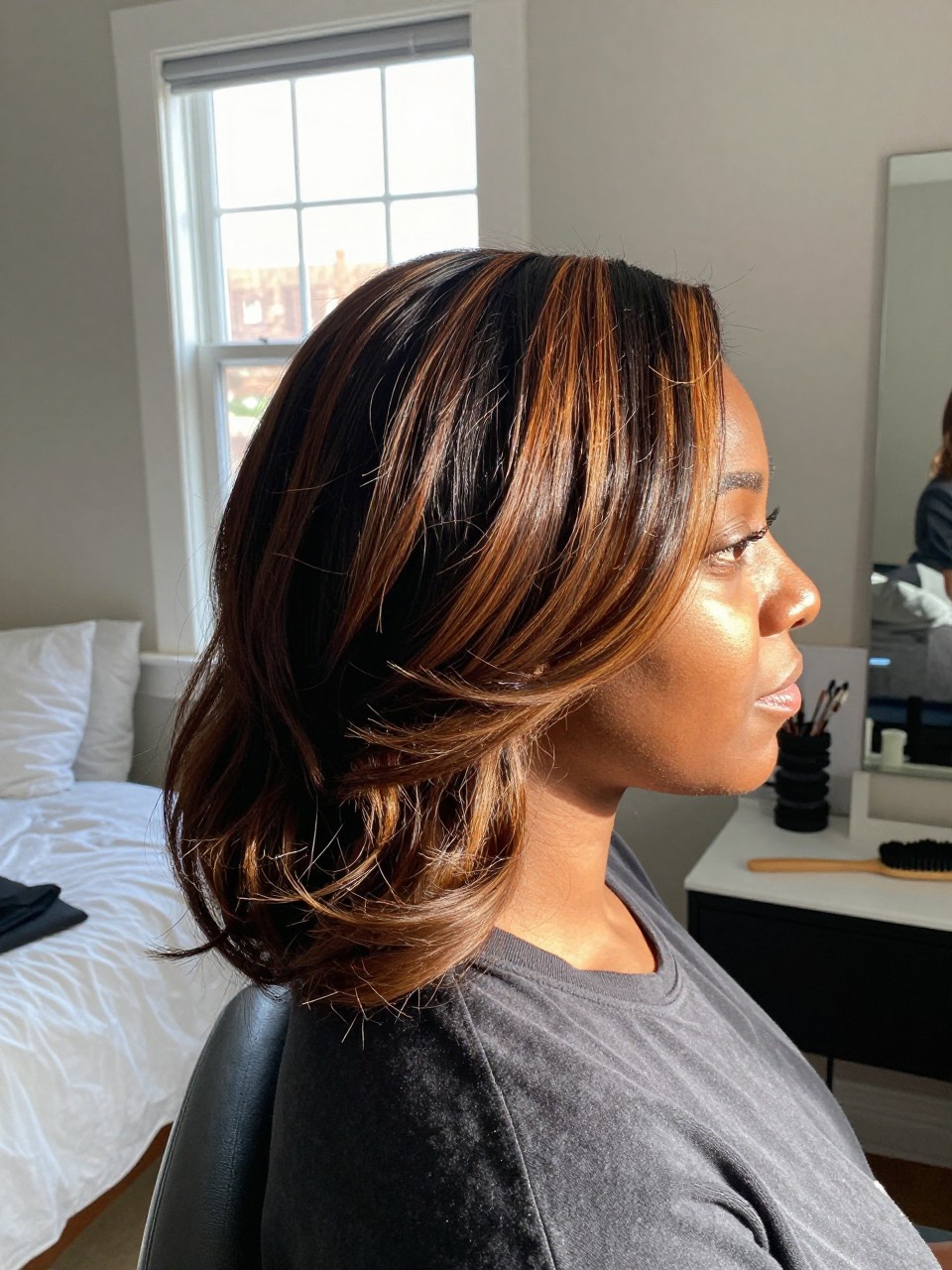 Photo of a Black woman wearing a side part dark blowout with maple honey brown highlights, side profile view, in a sun-drenched bedroom, natural window light, containing a small lived-in detail such as velcro rollers on the vanity and a round brush nearby, latest iPhone photo quality.