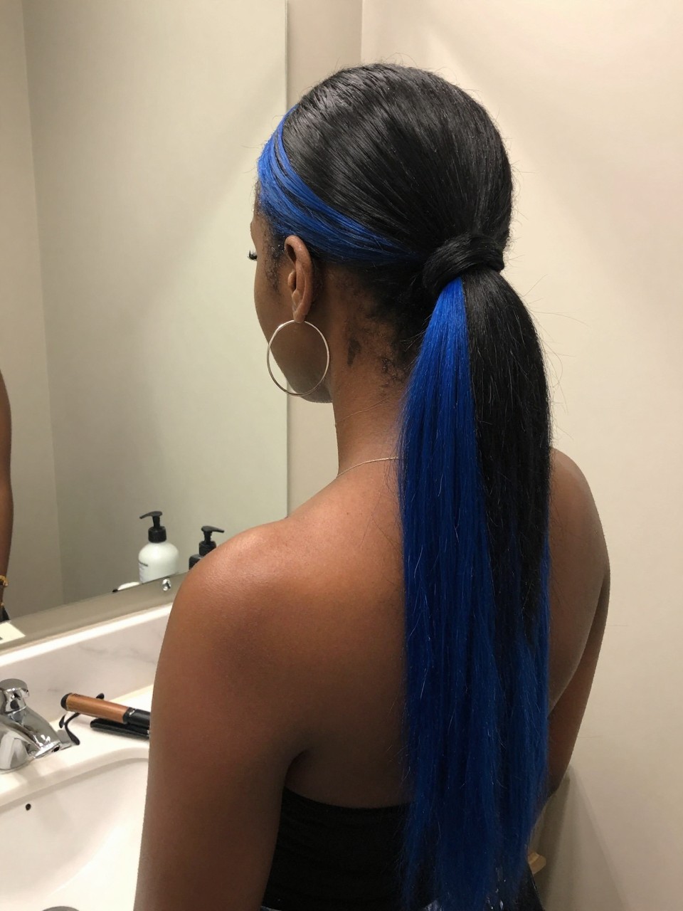 Photo of a black woman wearing a sleek mid ponytail with black hair and cobalt blue highlight panels through the tail, back-of-head view to highlight details, at a bathroom vanity mirror, soft indoor lighting, containing a small lived-in detail such as a flat iron on the counter and hoop earrings peeking through, latest iPhone photo quality.