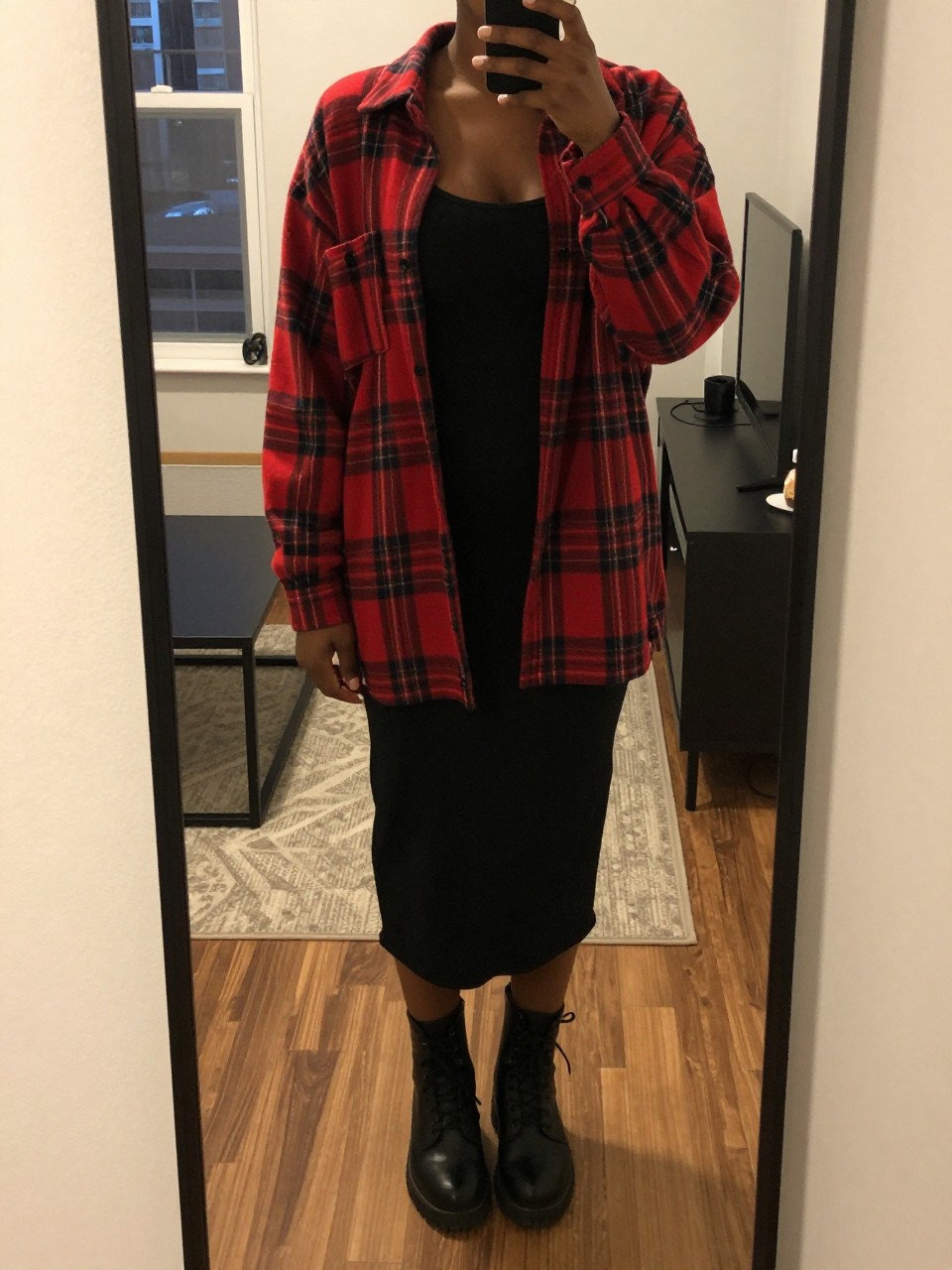 mirror selfie of a black woman wearing an oversized red plaid flannel open over a black tank top with a black midi skirt and black combat boots, one hand in her pocket, cropped at NECK so face doesn't show, in a city apartment, warm moody lighting, iPhone photo quality.