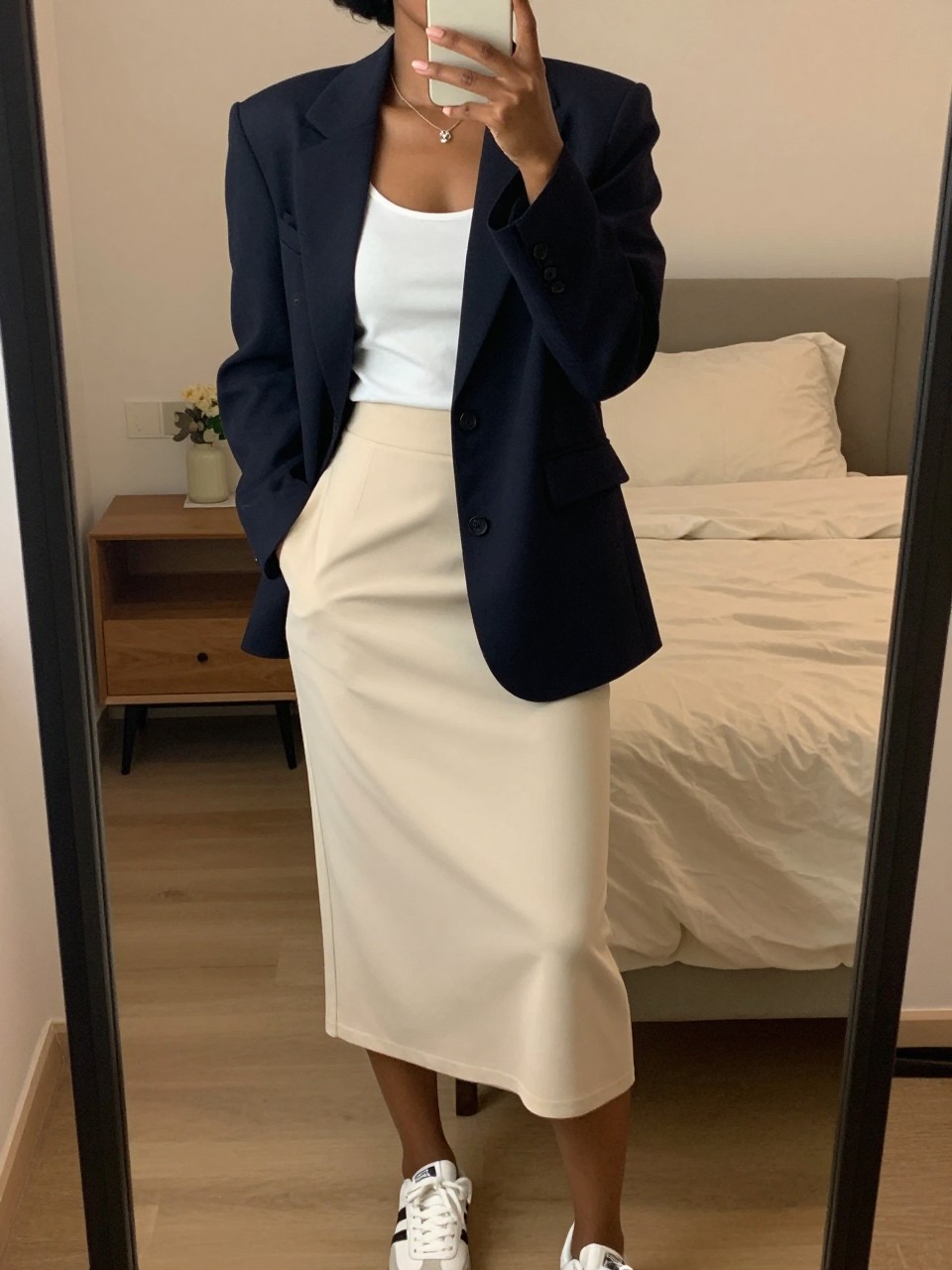 iPhone mirror selfie of a black woman wearing a navy blazer over a white tank top tucked into a soft cream midi skirt with white sneakers, one hand in her blazer pocket, cropped at NECK so face doesn't show, in a feminine-themed bedroom, warm moody lighting, iPhone photo quality.