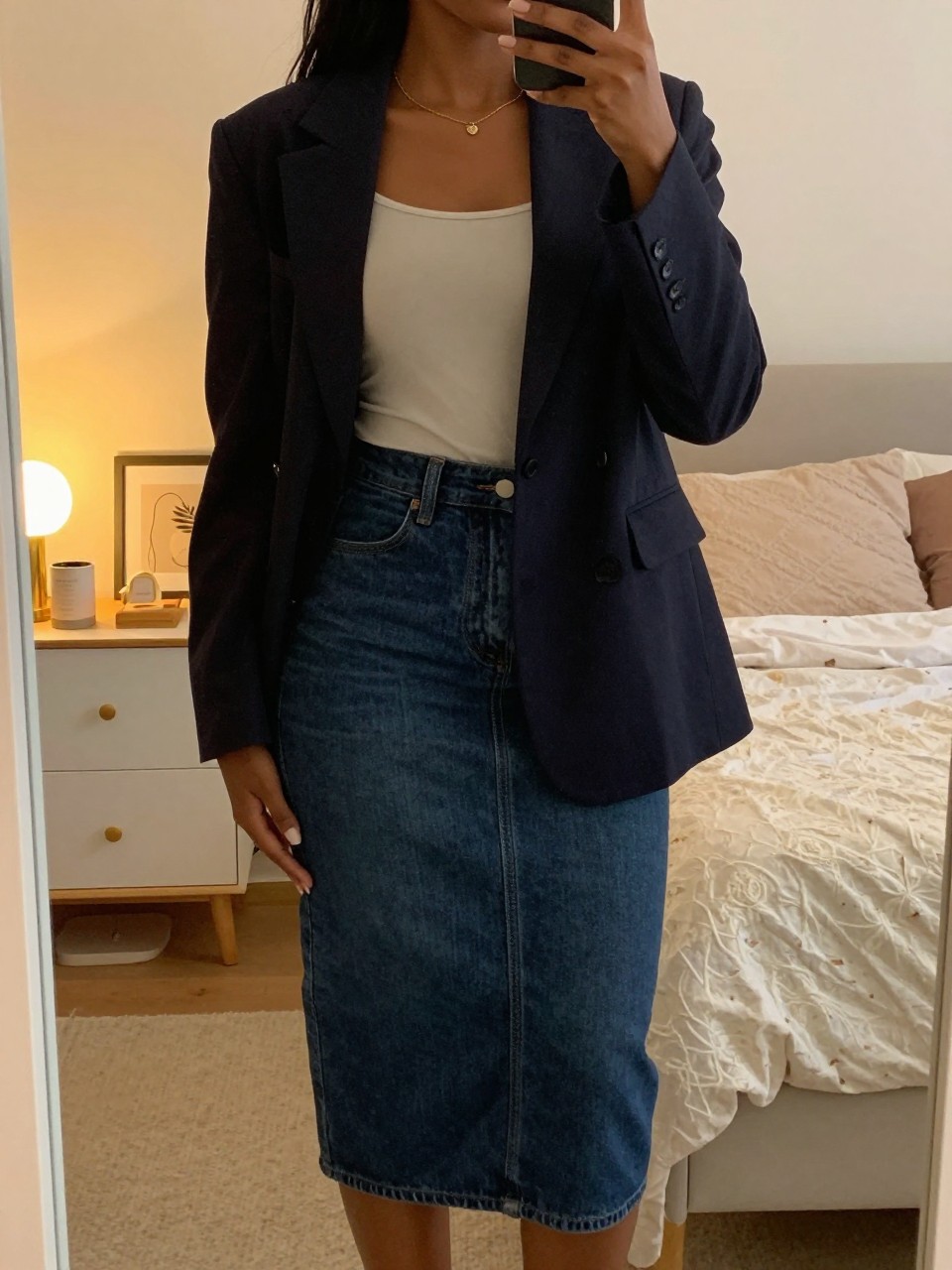 mirror selfie of a Black woman wearing a dark denim midi skirt with a blazer and simple top, standing with her weight shifted and phone at her side, cropped at NECK so face doesn't show, in a feminine-themed bedroom, evening golden hour light, iPhone photo quality.
