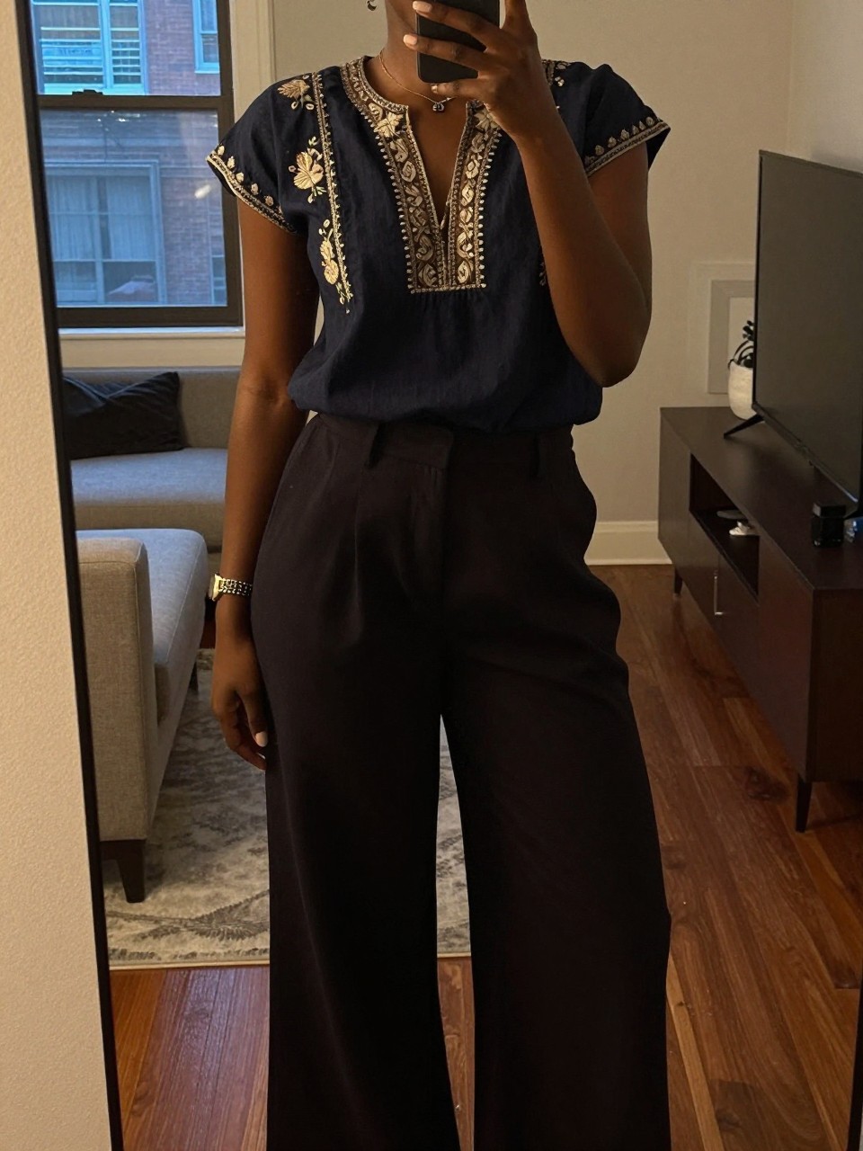 mirror selfie of a black woman in an embroidered top and wide-leg pants, showing her bracelet watch, cropped at NECK so face doesn't show, in a city apartment, warm moody lighting, iPhone photo quality.