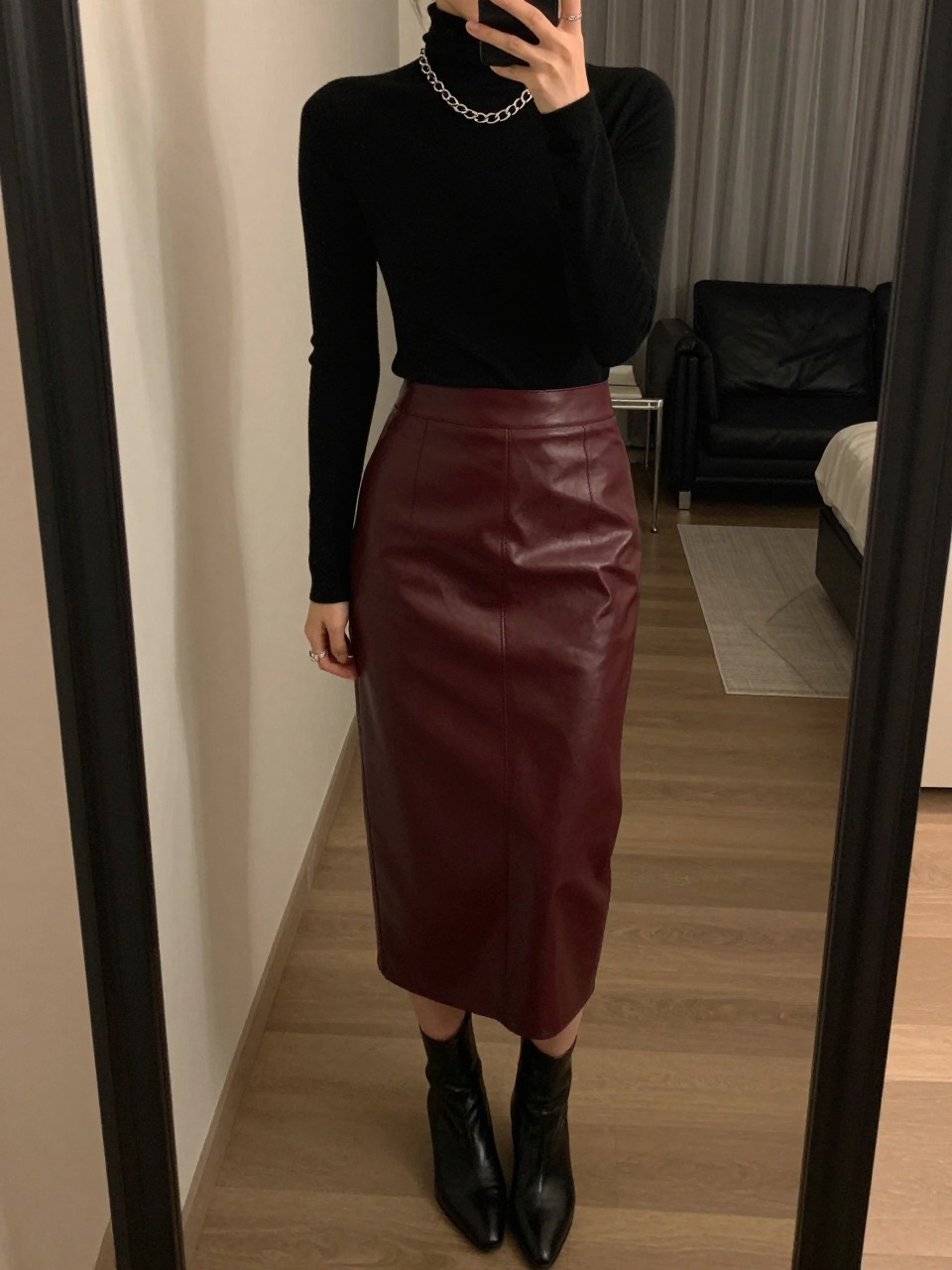 mirror selfie of a woman wearing a black turtleneck tucked into a deep burgundy leather midi skirt with a chunky silver chain necklace and black ankle boots, one hand touching her necklace, cropped at NECK so face doesn't show, in a city apartment, warm moody lighting, iPhone photo quality.