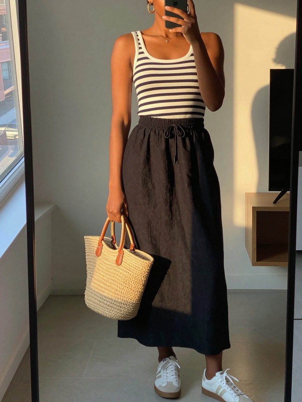 mirror selfie of a black woman wearing a navy and white striped boatneck top tucked into a black linen maxi skirt with white sneakers and gold hoop earrings, holding a straw tote bag, standing in a relaxed chic pose, cropped at NECK so face doesn't show, in a city apartment, evening golden hour, iPhone photo quality.