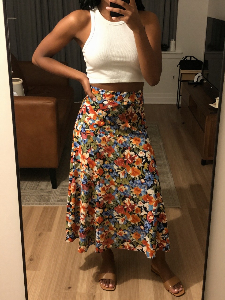 mirror selfie of a black woman wearing a white ribbed crop top with a high-waisted floral print flowy midi skirt and flat tan sandals, one hand on her hip, cropped at NECK so face doesn't show, in a city apartment, warm moody lighting, iPhone photo quality.