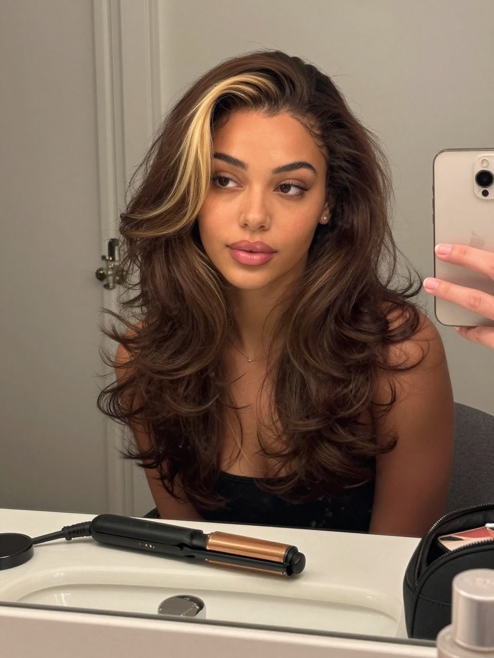 Photo of a biracial woman wearing brunette hair with a vanilla blonde money piece and soft face-framing waves, candid mirror selfie, bathroom vanity mirror setting, soft indoor lighting, with a flat iron on the counter and a makeup bag nearby, latest iPhone photo quality.