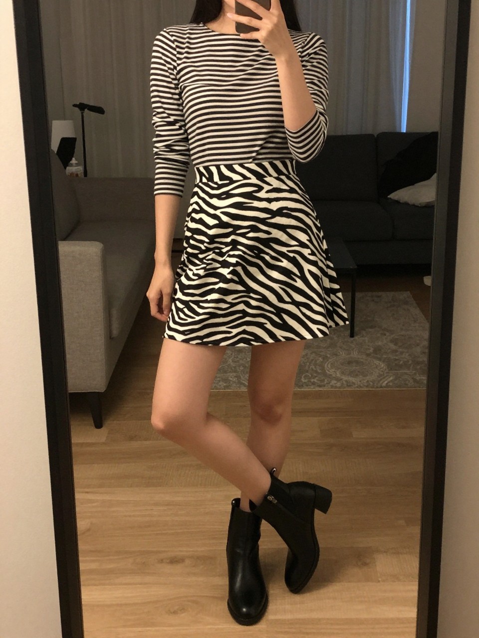 iPhone mirror selfie of a woman in a striped top and a zebra print A-line skirt with ankle boots, standing with her weight on one leg, cropped at NECK so face doesn't show, in a city apartment, warm moody lighting, iPhone photo quality.