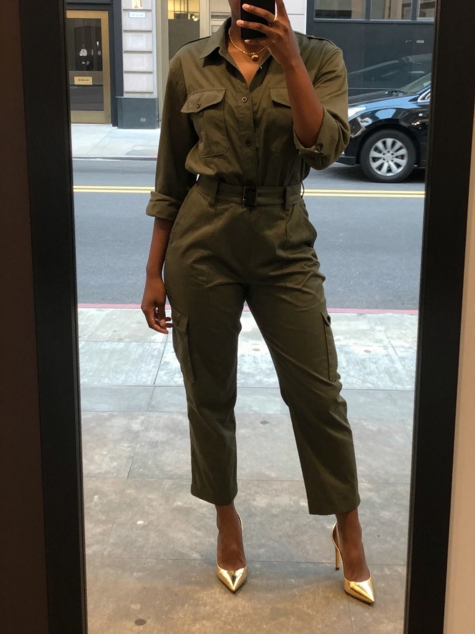 mirror selfie of a black woman in a green utility jumpsuit and gold heels, standing in a city street with cool architecture, cropped at NECK so face doesn't show, in a city apartment, warm moody lighting, iPhone photo quality.