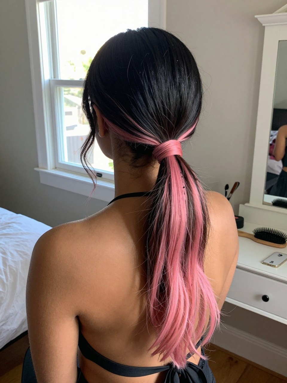 Photo of a biracial woman wearing a sleek low ponytail with black hair and pink highlights through the tail and ends, back-of-head view to highlight details, in a sun-drenched bedroom, natural window light, containing a small lived-in detail such as a black ribbon tied at the base and a hairbrush on the vanity, latest iPhone photo quality.