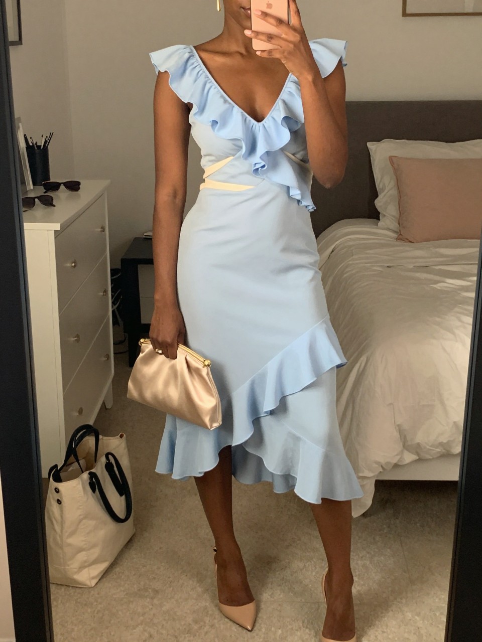 mirror selfie of a black woman wearing a soft sky blue ruffle midi dress with a cream light wrap, nude slingback heels, simple gold drop earrings, and a champagne satin clutch bag, holding a phone in front of a mirror, cropped at NECK so face doesn't show, with a natural casual selfie pose, in a feminine-themed bedroom, warm moody lighting, including small lived-in details like a tote bag on the floor, sunglasses on a dresser, or soft bedding in the background, latest iPhone photo quality.