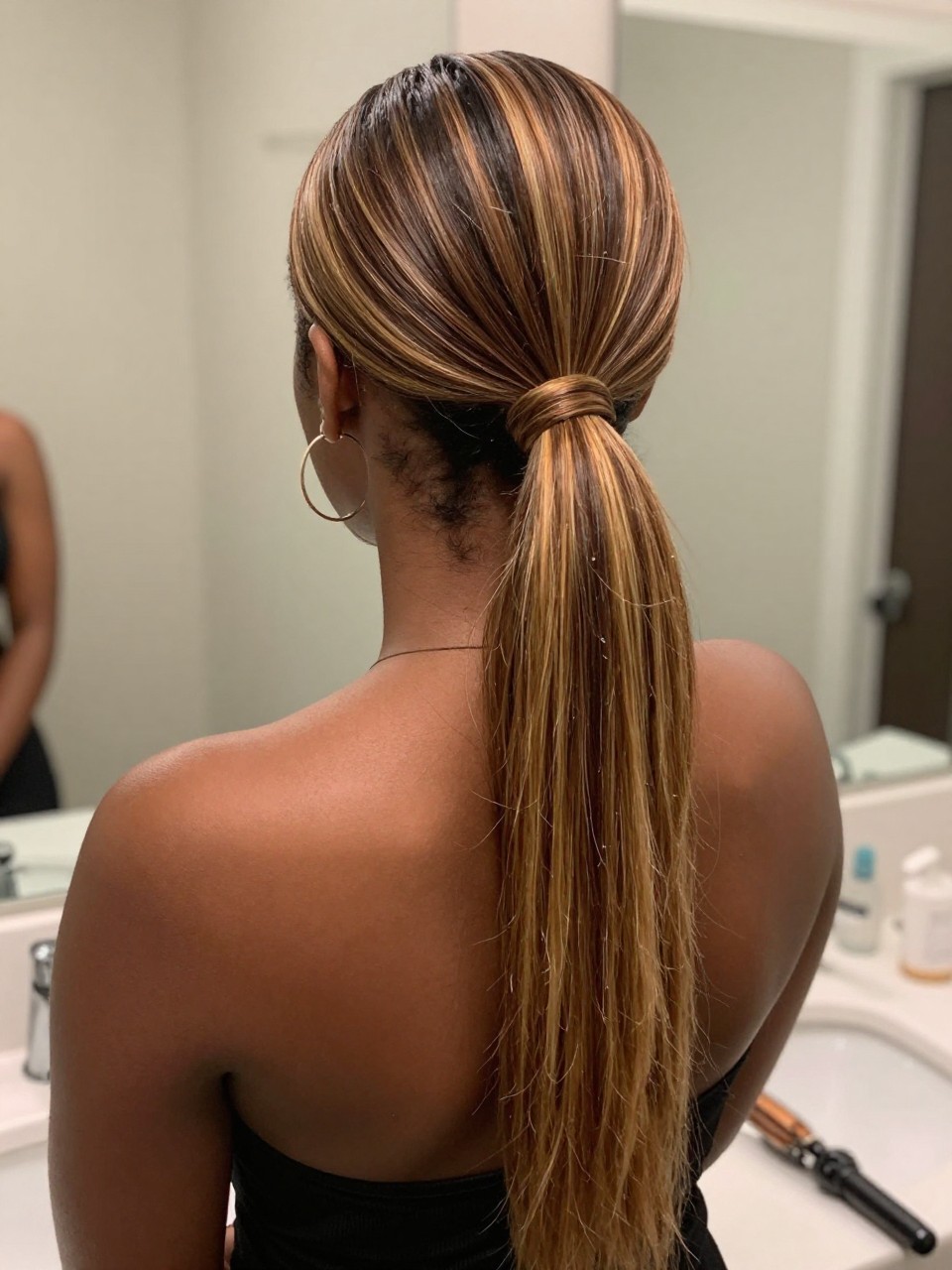 Photo of a black woman wearing a sleek mid ponytail with brown hair and golden chunky highlight streaks through the tail, back-of-head view to highlight details, at a bathroom vanity mirror, soft indoor lighting, containing a small lived-in detail such as a flat iron on the counter and hoop earrings peeking through, latest iPhone photo quality.