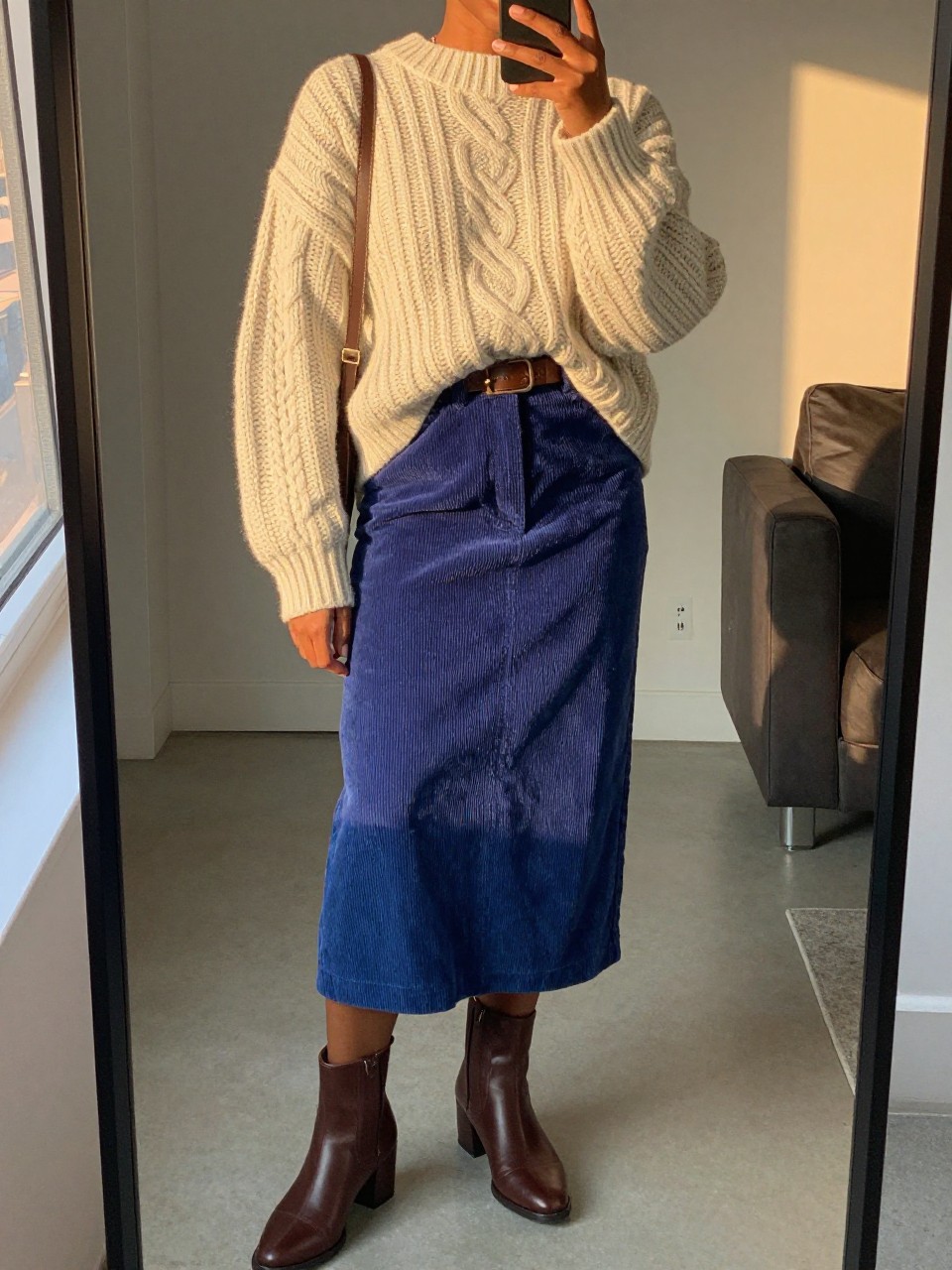 mirror selfie of a black woman wearing an oversized cream chunky cable-knit sweater loosely tucked into a cobalt blue corduroy midi skirt with a brown leather belt and matching brown block-heel ankle boots, holding a brown crossbody bag, standing in a cozy pose, cropped at NECK so face doesn't show, in a city apartment, evening golden hour, iPhone photo quality.