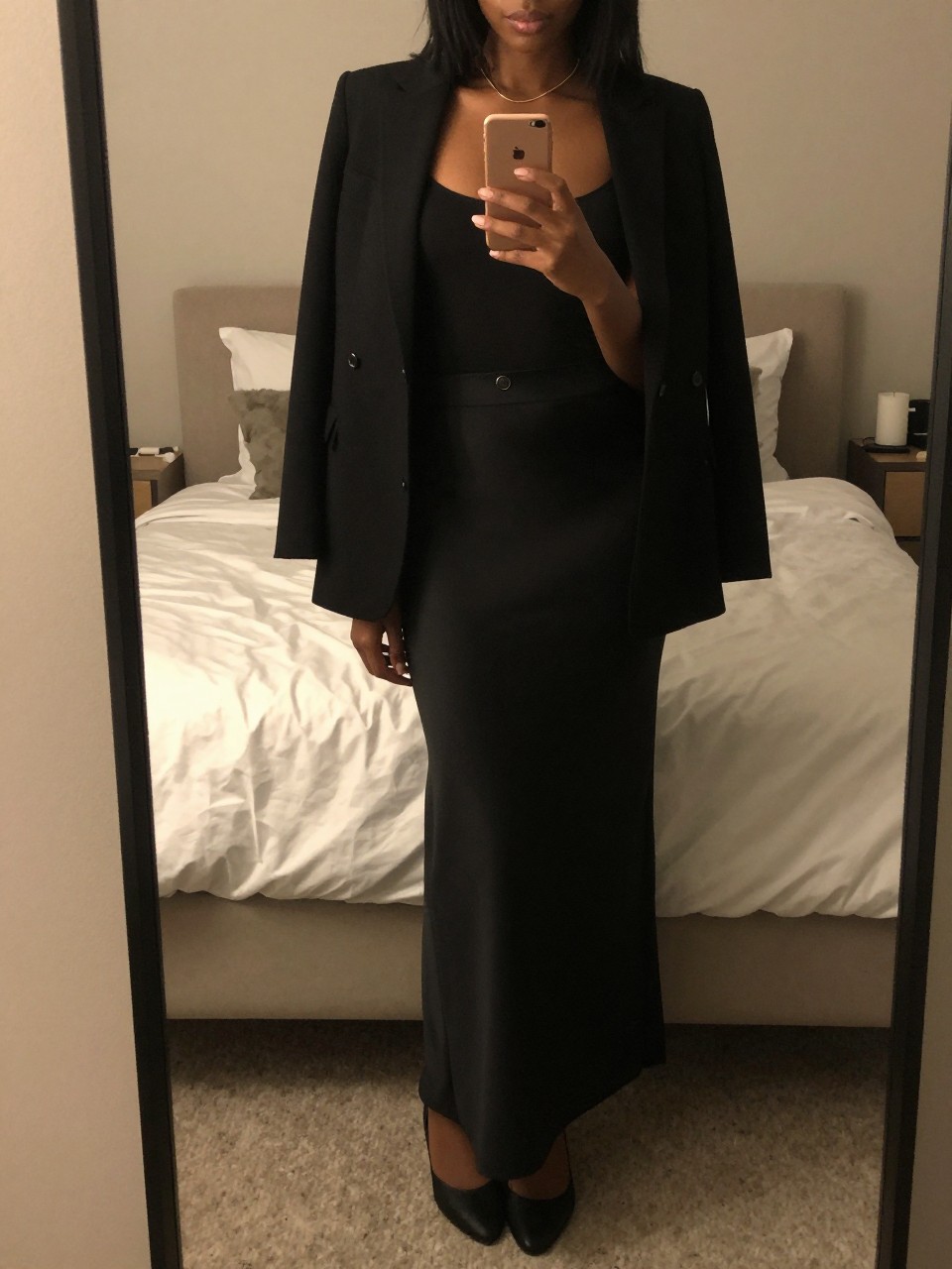 iPhone mirror selfie of a black woman in a black maxi skirt and blazer over a bodysuit with flats, holding phone at chest, cropped at NECK so face doesn't show, in a feminine-themed bedroom, warm moody lighting, iPhone photo quality.