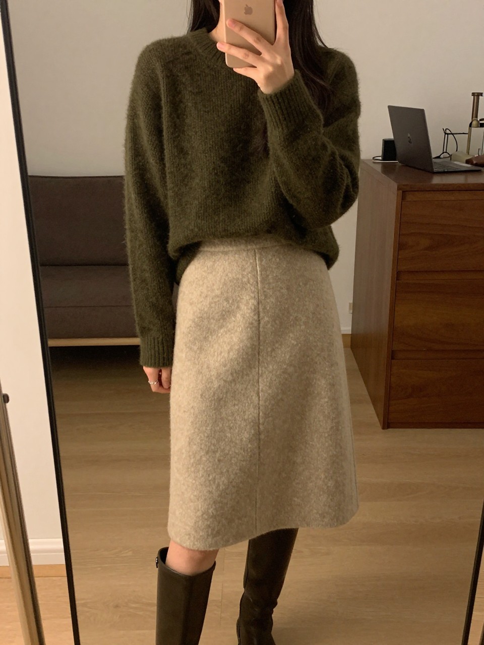 mirror selfie of a woman in an olive green sweater and a beige wool skirt with knee-high boots, standing with phone in hand, cropped at NECK so face doesn't show, in a city apartment, warm moody lighting, iPhone photo quality.