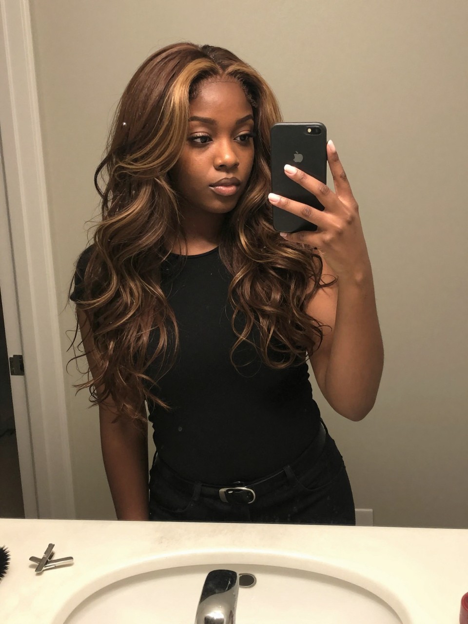 Photo of a black woman wearing a wavy brown lob with ash blonde chunky highlights, candid mirror selfie, at a bathroom vanity mirror, soft indoor lighting, containing a small lived-in detail such as a hairbrush on the sink and a silver clip nearby, latest iPhone photo quality.
