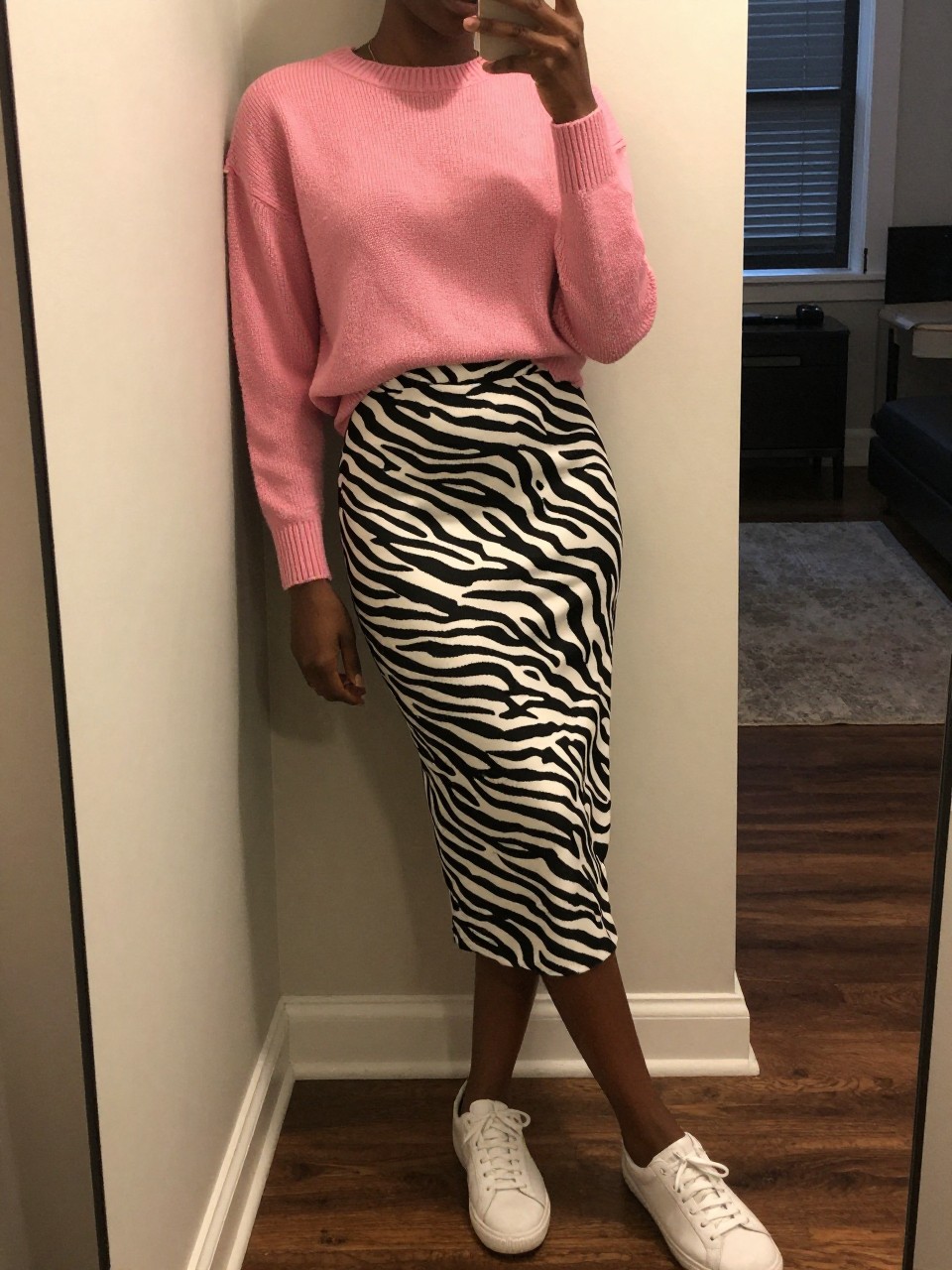 mirror selfie of a black woman in a pink sweater and a zebra print midi skirt with white sneakers, casually leaning against a wall, cropped at NECK so face doesn't show, in a city apartment, warm moody lighting, iPhone photo quality.