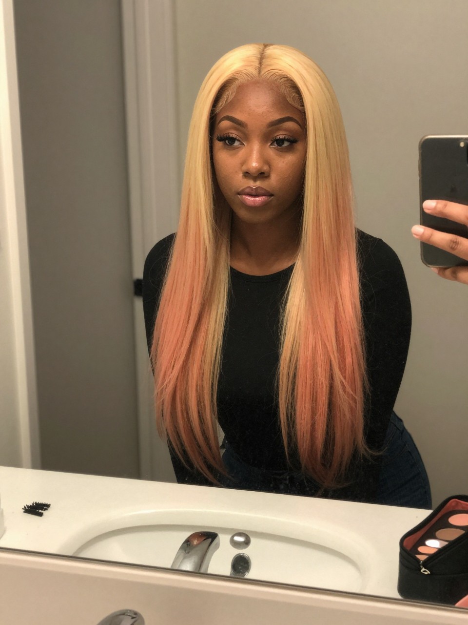 Photo of a black woman wearing strawberry blonde curtain layers with golden peach highlights around the face, candid mirror selfie, at a bathroom vanity mirror, soft indoor lighting, containing a small lived-in detail such as mini sectioning clips on the counter and a makeup pouch nearby, latest iPhone photo quality.