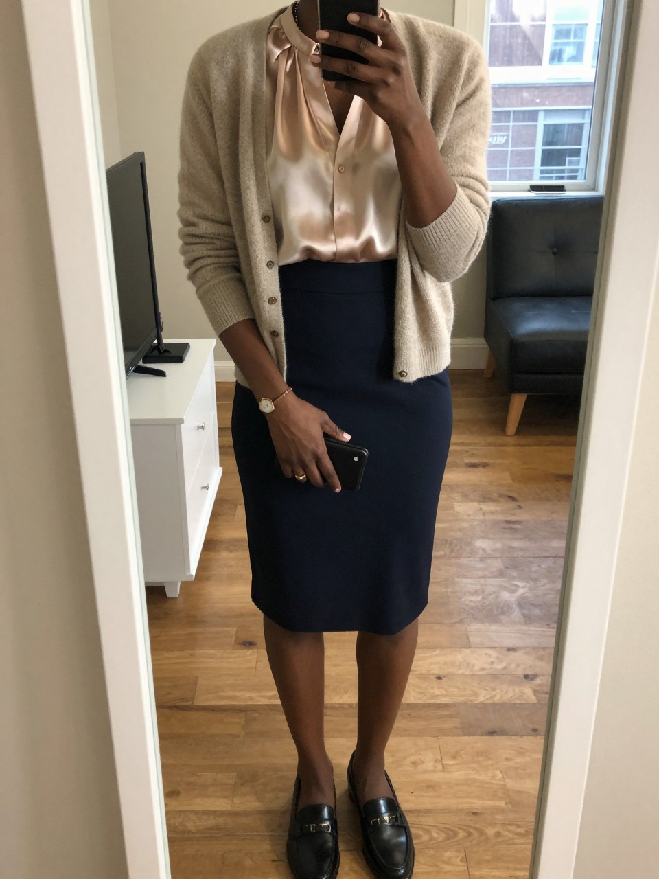 iPhone mirror selfie of a black woman wearing a cashmere cardigan over a silk shell tucked into a navy pencil skirt with loafers, her hands clasped in front holding a small wallet, cropped at NECK so face doesn't show, in a bright city apartment, warm moody lighting, iPhone photo quality.