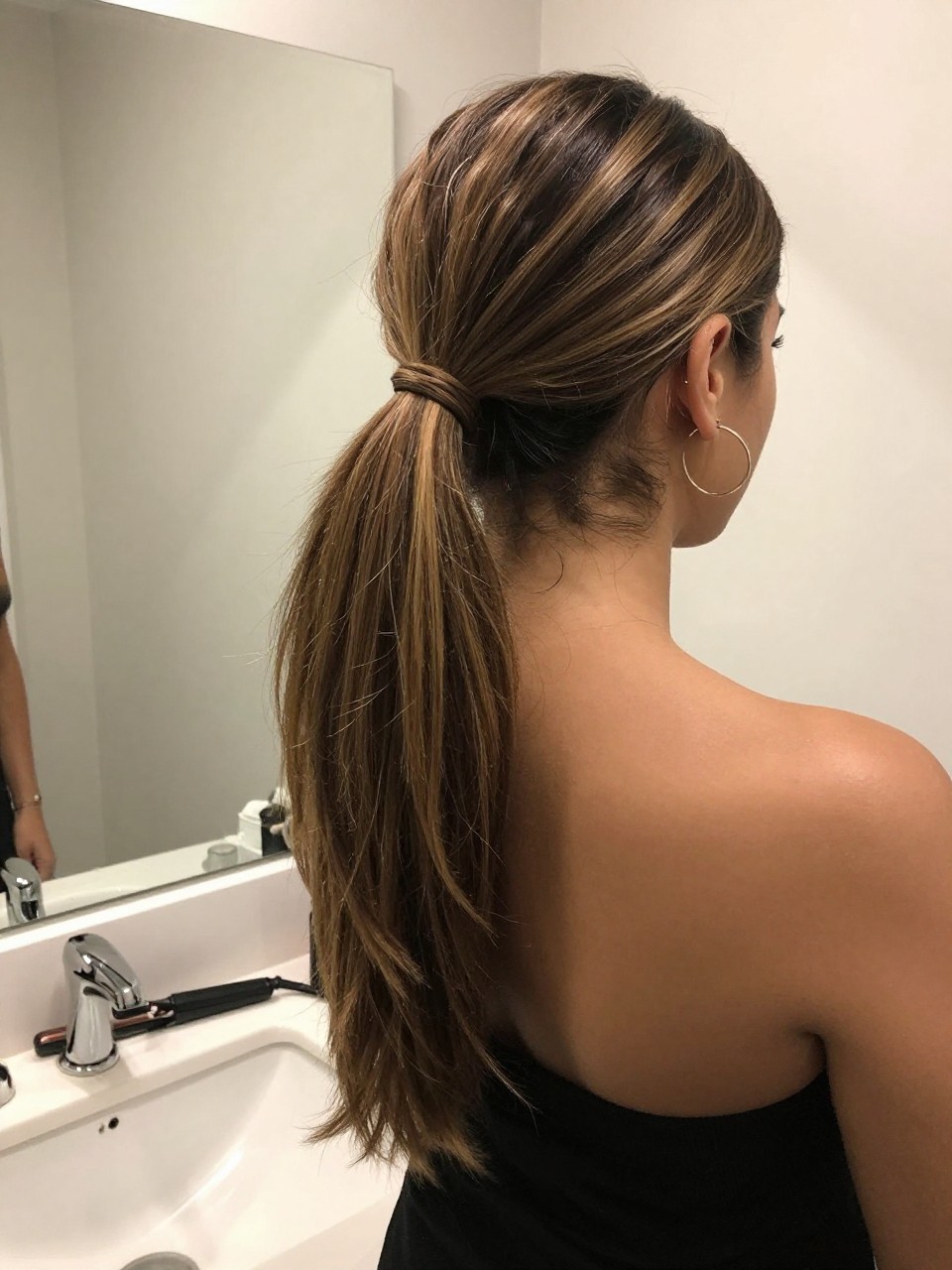 Photo of a woman wearing a sleek mid ponytail with mixed brunette highlights and lowlights through the lengths, back-of-head view to highlight details, at a bathroom vanity mirror, soft indoor lighting, containing a small lived-in detail such as a flat iron on the counter and hoop earrings peeking through, latest iPhone photo quality.
