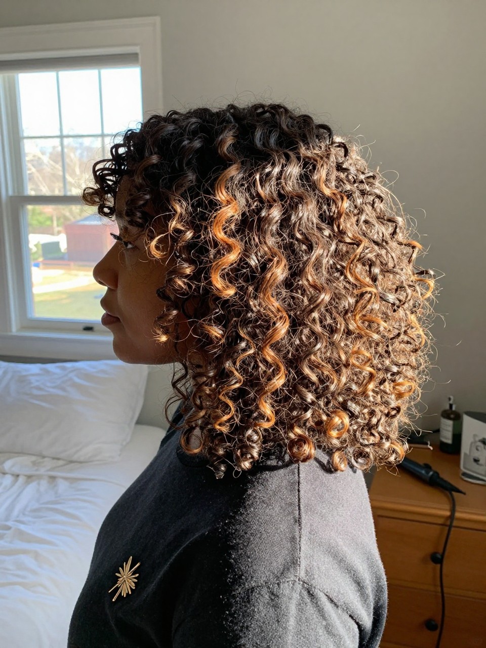 Photo of a black woman wearing soft dark curls with caramel honey highlights, side profile view, in a sun-drenched bedroom, natural window light, containing a small lived-in detail such as a decorative pin on one side and a curling iron resting on the nightstand, latest iPhone photo quality.