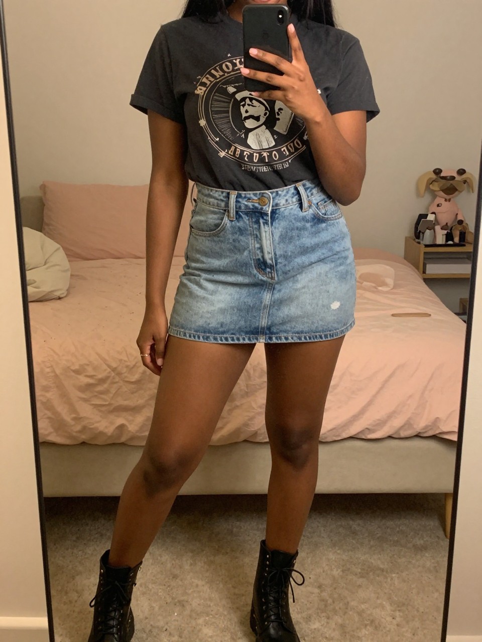mirror selfie of a Black woman wearing a light wash denim mini skirt with a graphic tee and combat boots, posed with her phone low and one foot forward, cropped at NECK so face doesn't show, in a feminine-themed bedroom, warm moody lighting, iPhone photo quality.