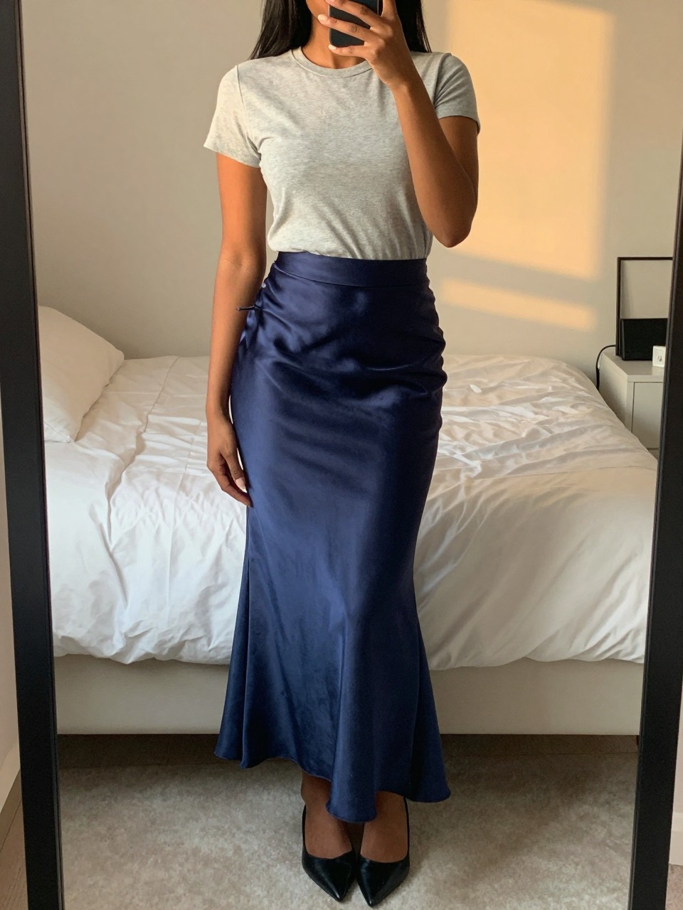 mirror selfie of a black woman in a grey t-shirt and a navy satin mermaid skirt with pointed flats, gently touching her hair, cropped at NECK so face doesn't show, in a feminine-themed bedroom, evening golden hour, iPhone photo quality.