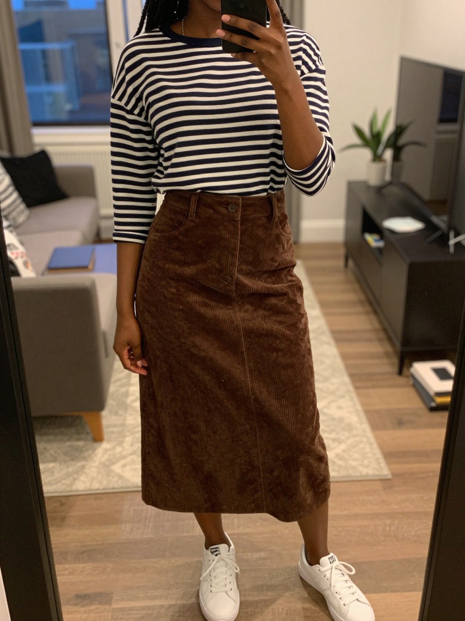 iPhone mirror selfie of a black woman wearing a navy striped Breton top tucked into a chocolate brown A-line corduroy skirt with white sneakers, one hand holding her phone at her side, cropped at NECK so face doesn't show, in a city apartment, warm moody lighting, iPhone photo quality.